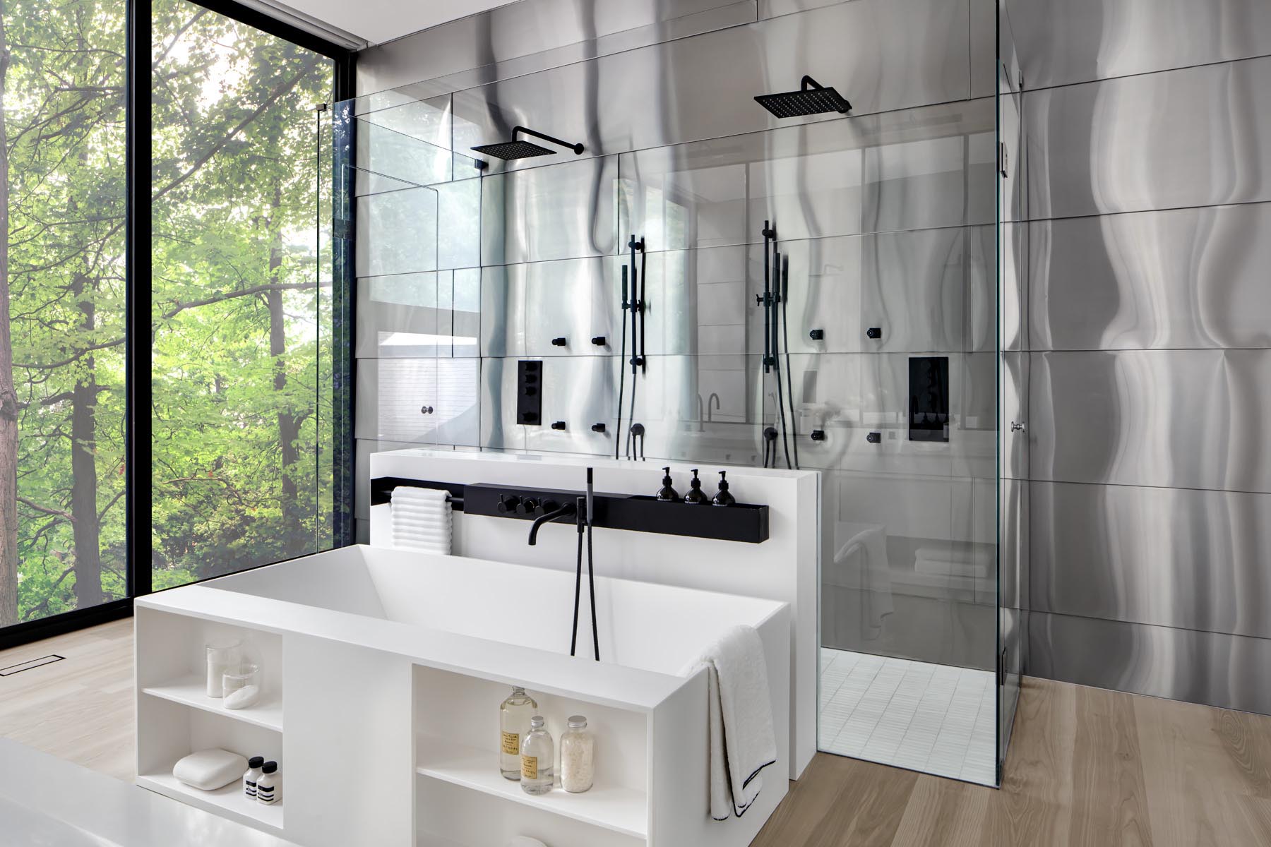 A wall of wood closets link the bedroom to this modern en-suite bathroom, that showcases a white vanity with large mirrors, a built-in bathtub, and a double shower with glass surround.