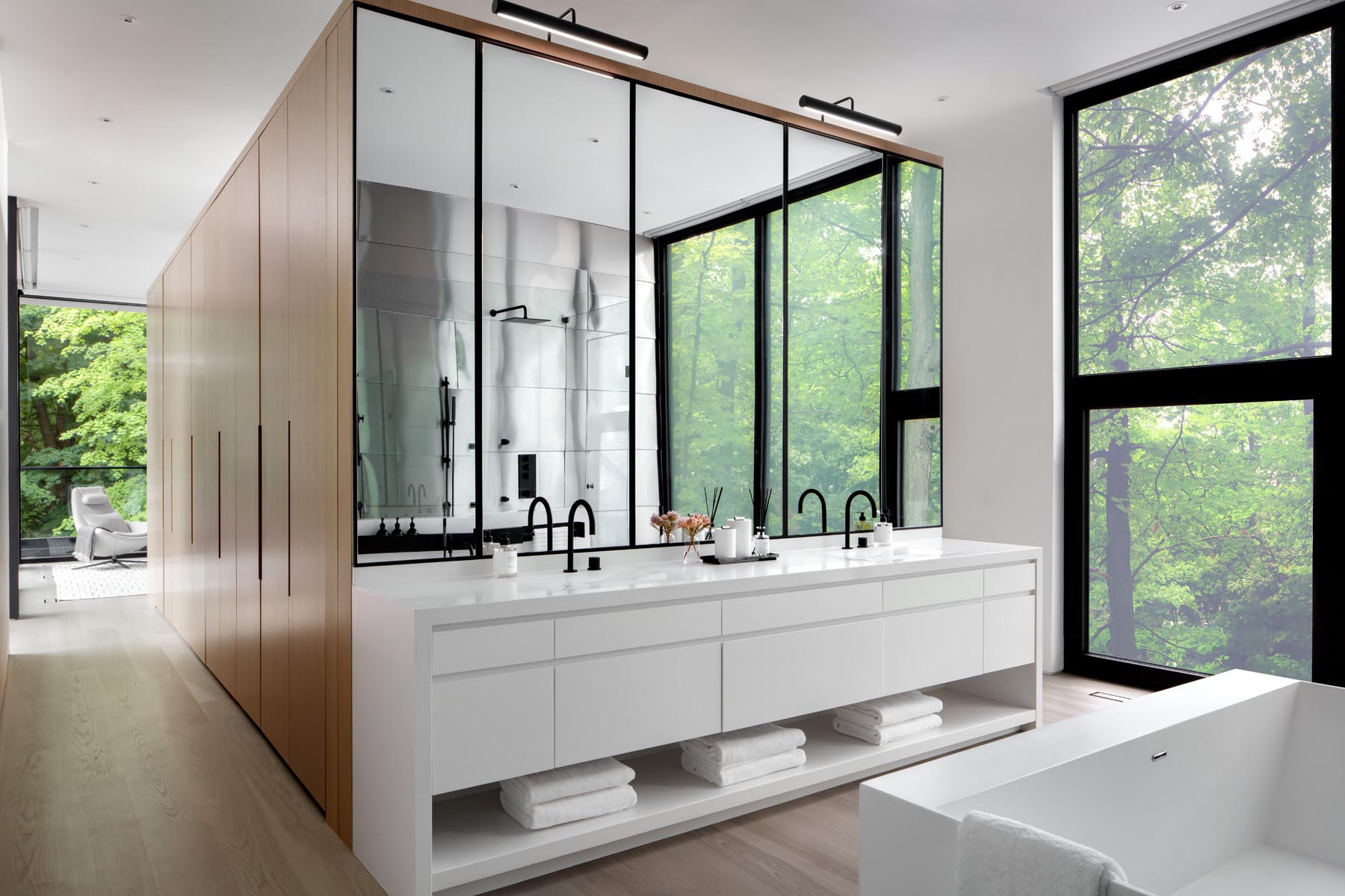 A wall of wood closets link the bedroom to this modern en-suite bathroom, that showcases a white vanity with large mirrors, a built-in bathtub, and a double shower with glass surround.