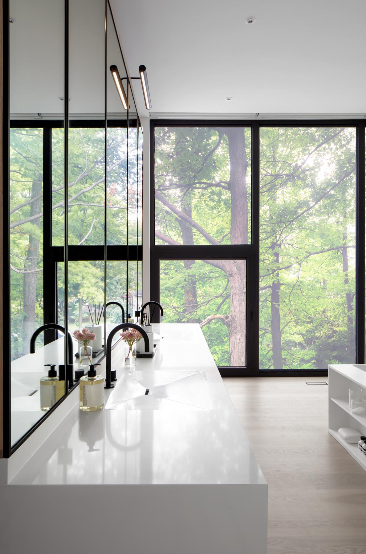 A wall of wood closets link the bedroom to this modern en-suite bathroom, that showcases a white vanity with large mirrors, a built-in bathtub, and a double shower with glass surround.