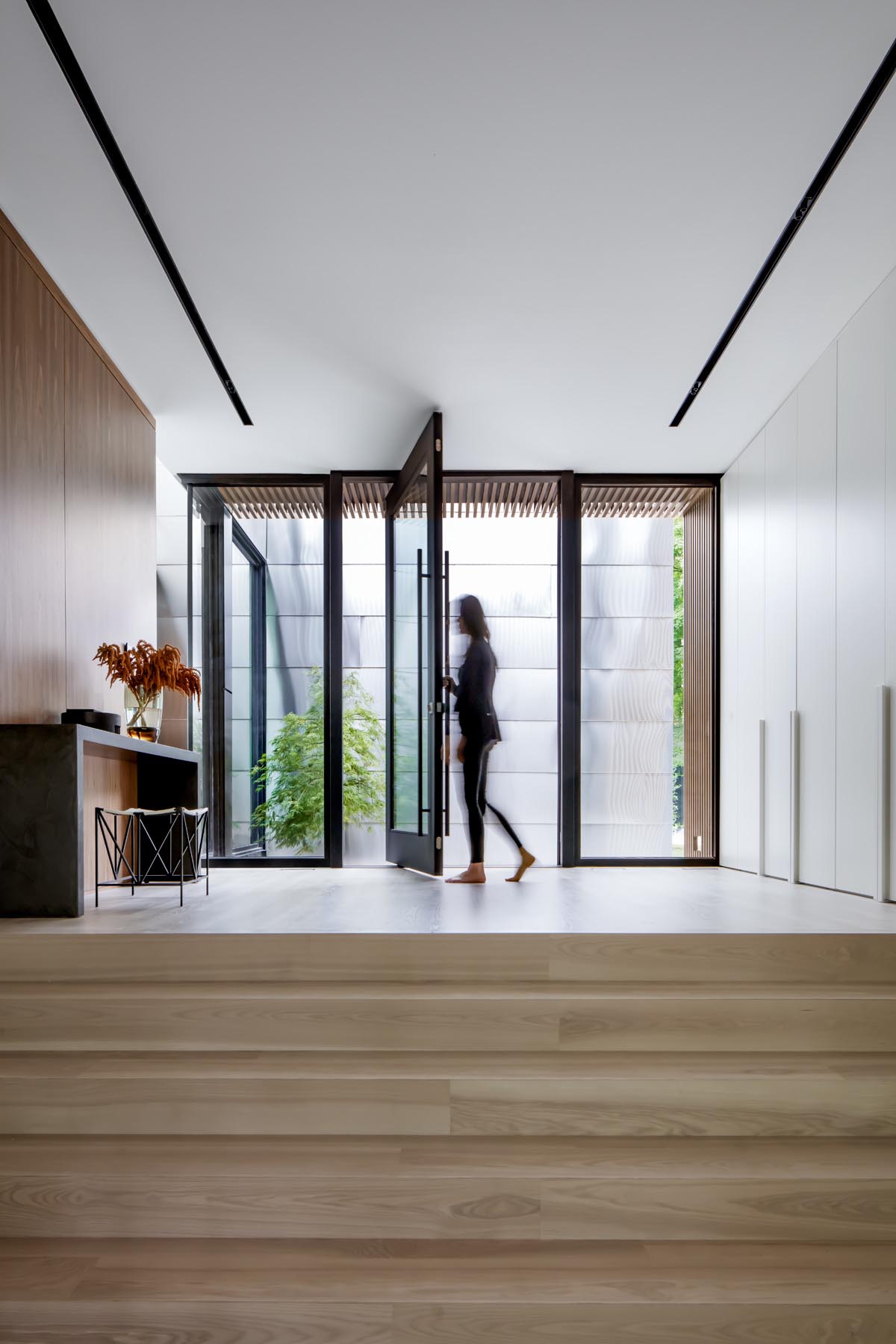 Welcoming visitors to this modern home is a 7-foot wide black-framed glass pivot door, that opens into the entryway furnished with a console table and a stool. 