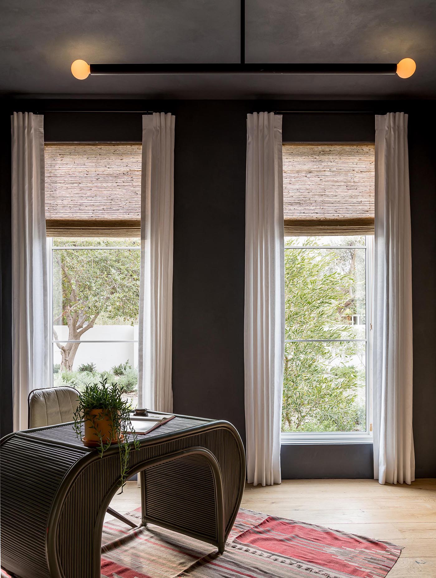 This modern home office has a dramatic appearance, with dark gray walls, a minimalist light fixture, tall white curtains, a curved desk, and colorful rug.