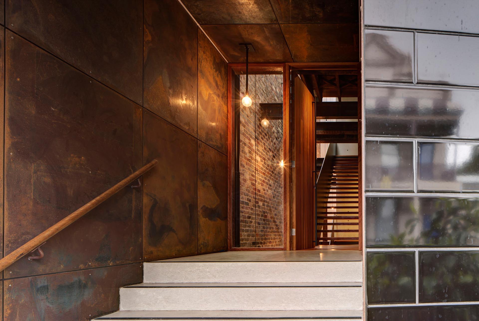 Stairs and a wall covered with aged steel lead the way to the tall wood front door of this modern home.