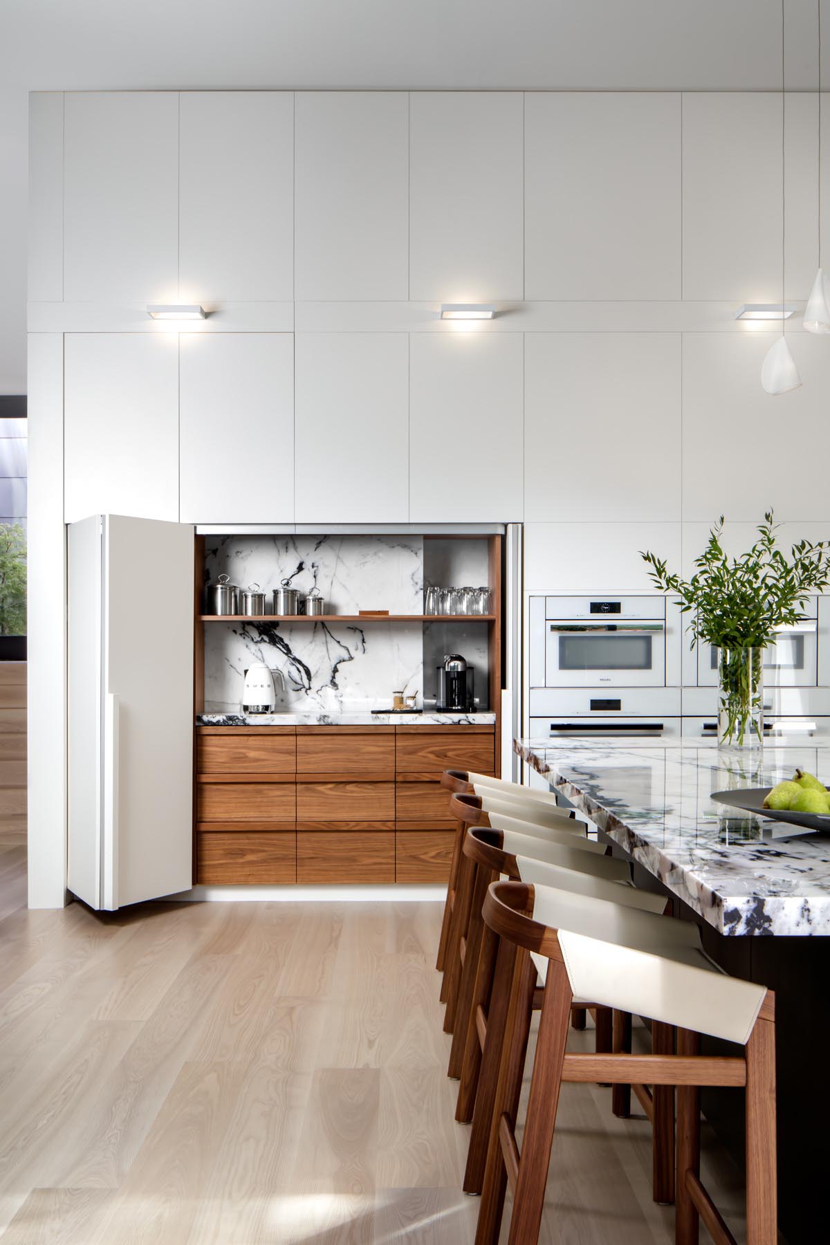 In this kitchen, minimalist white cabinets line the walls, while a large island provides space for multiple stools. Hidden within the cabinetry is a dedicated coffee station.