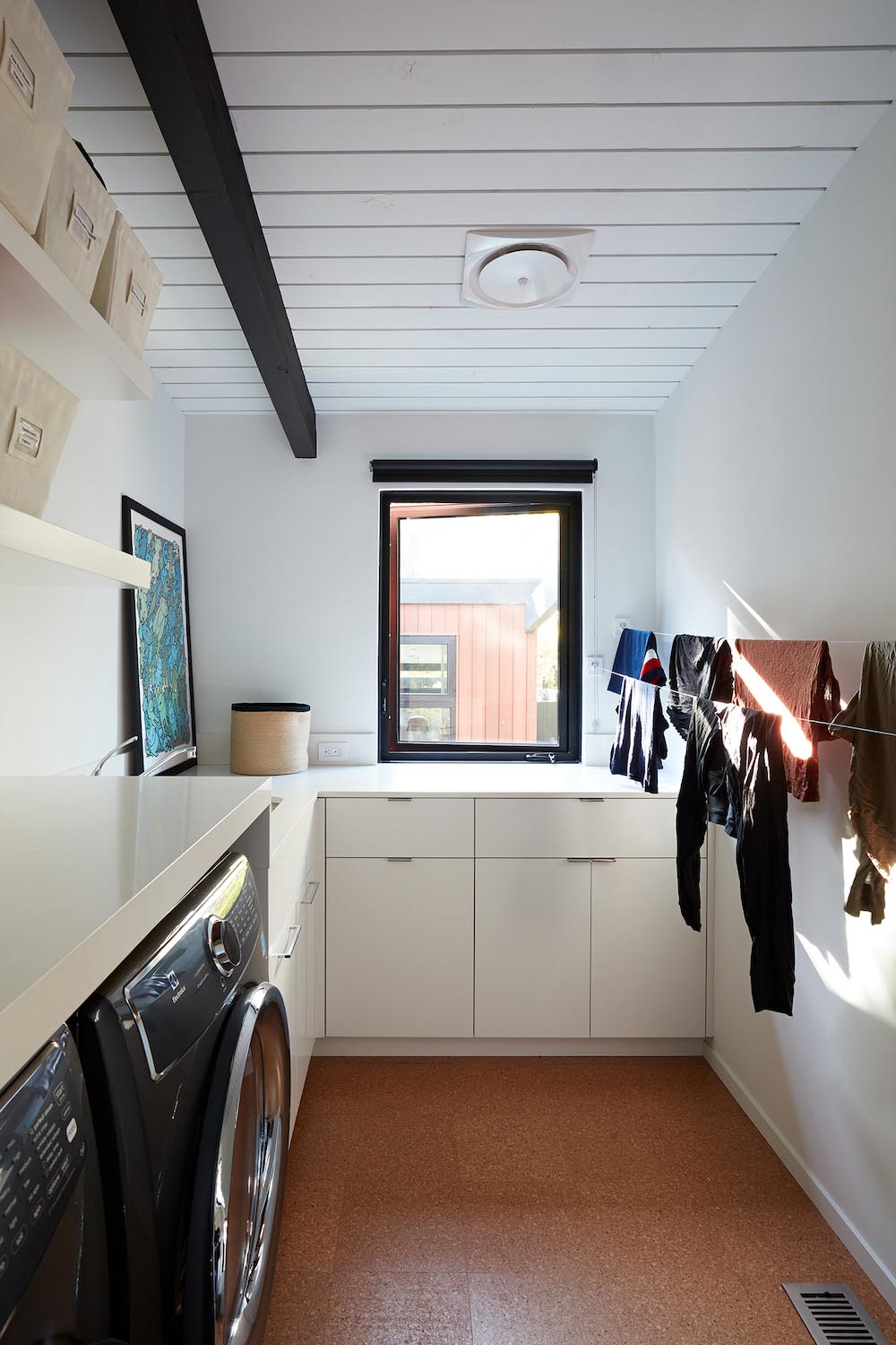 This modern laundry room includes cabinetry that wraps around the corner of the room, a countertop that continues above the washer and dryer, floating shelves, and drying lines.