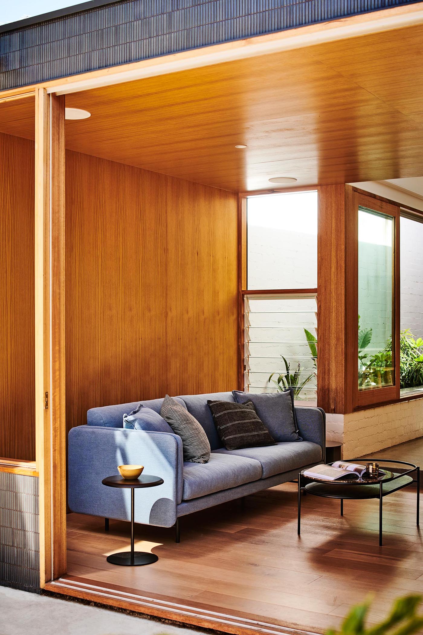 A modern sitting area, which opens to the backyard, includes wood walls, louver windows, shelving, and a colorful blue couch.