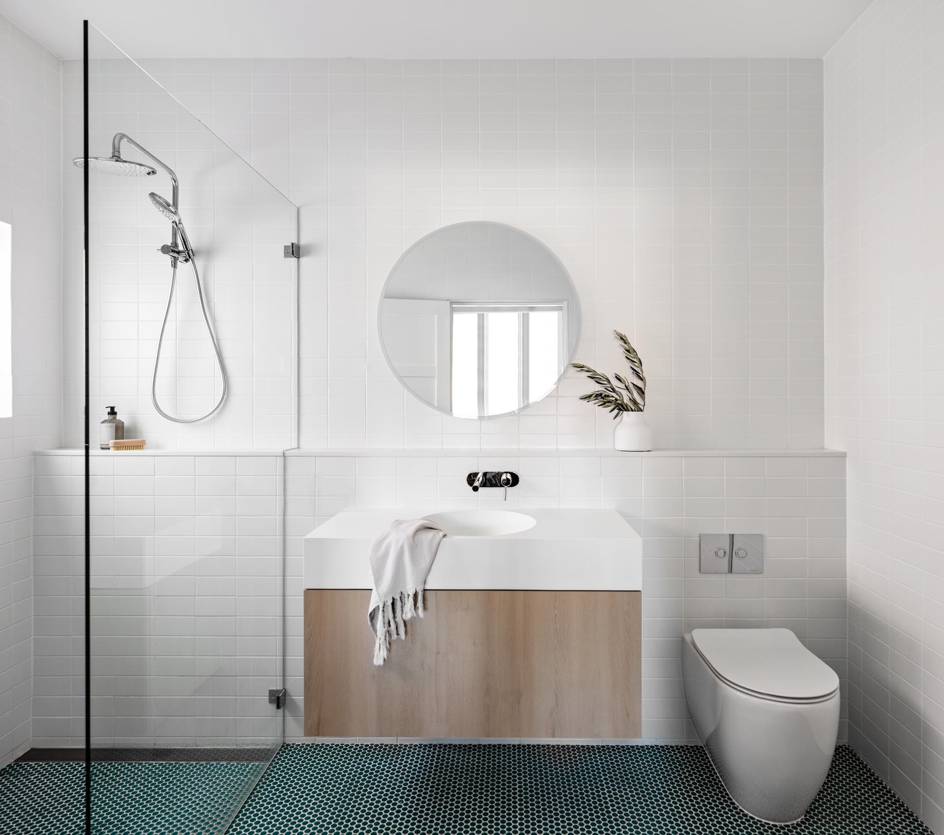 A modern bathroom with white wall tiles, white grout, a round mirror, a wood vanity with thick white countertop, and dark green penny tile flooring.