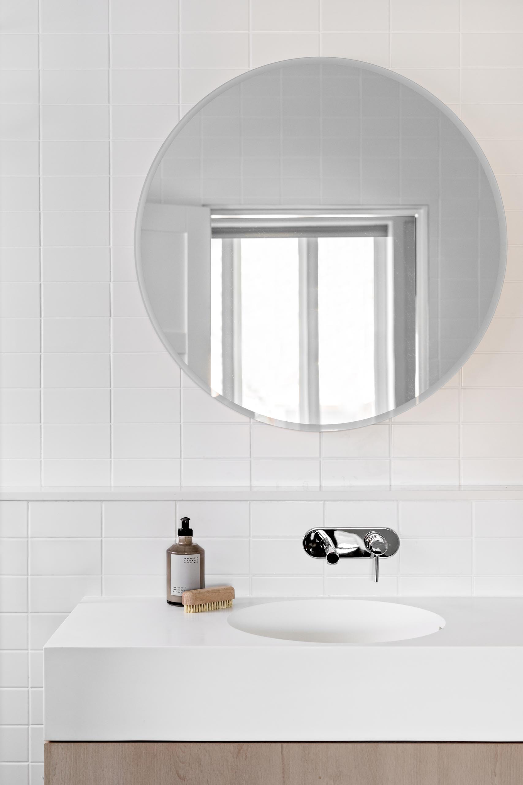 A modern white bathroom with white tiles, white grout, a round mirror, and wood vanity with thick white countertop.