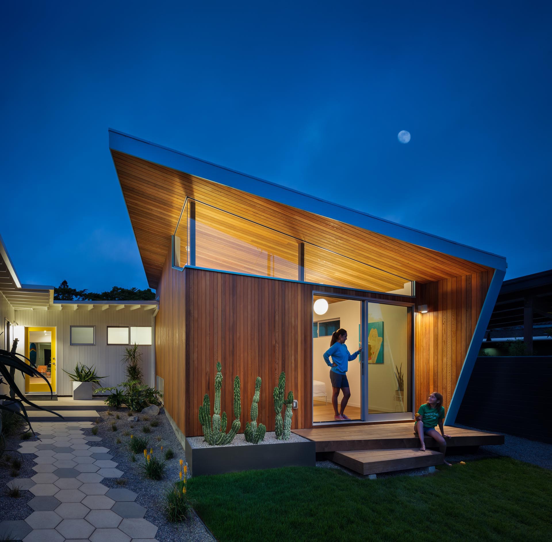 The updated exterior of this mid-century modern home showcases more usable green space and a permeable grass driveway, while an all new pavilion stands out by the tall-angled roofline with blue accents, and hexagonal pavers lead to the yellow front door.