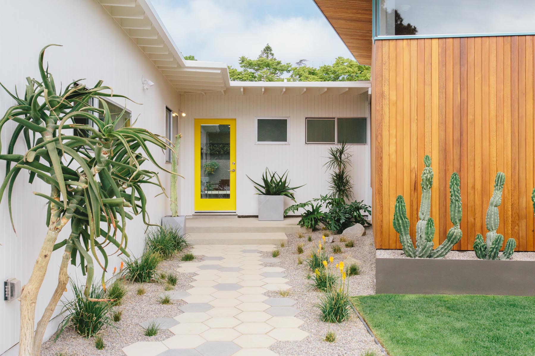 The updated exterior of this mid-century modern home showcases more usable green space and a permeable grass driveway, while an all new pavilion stands out by the tall-angled roofline with blue accents, and hexagonal pavers lead to the yellow front door.