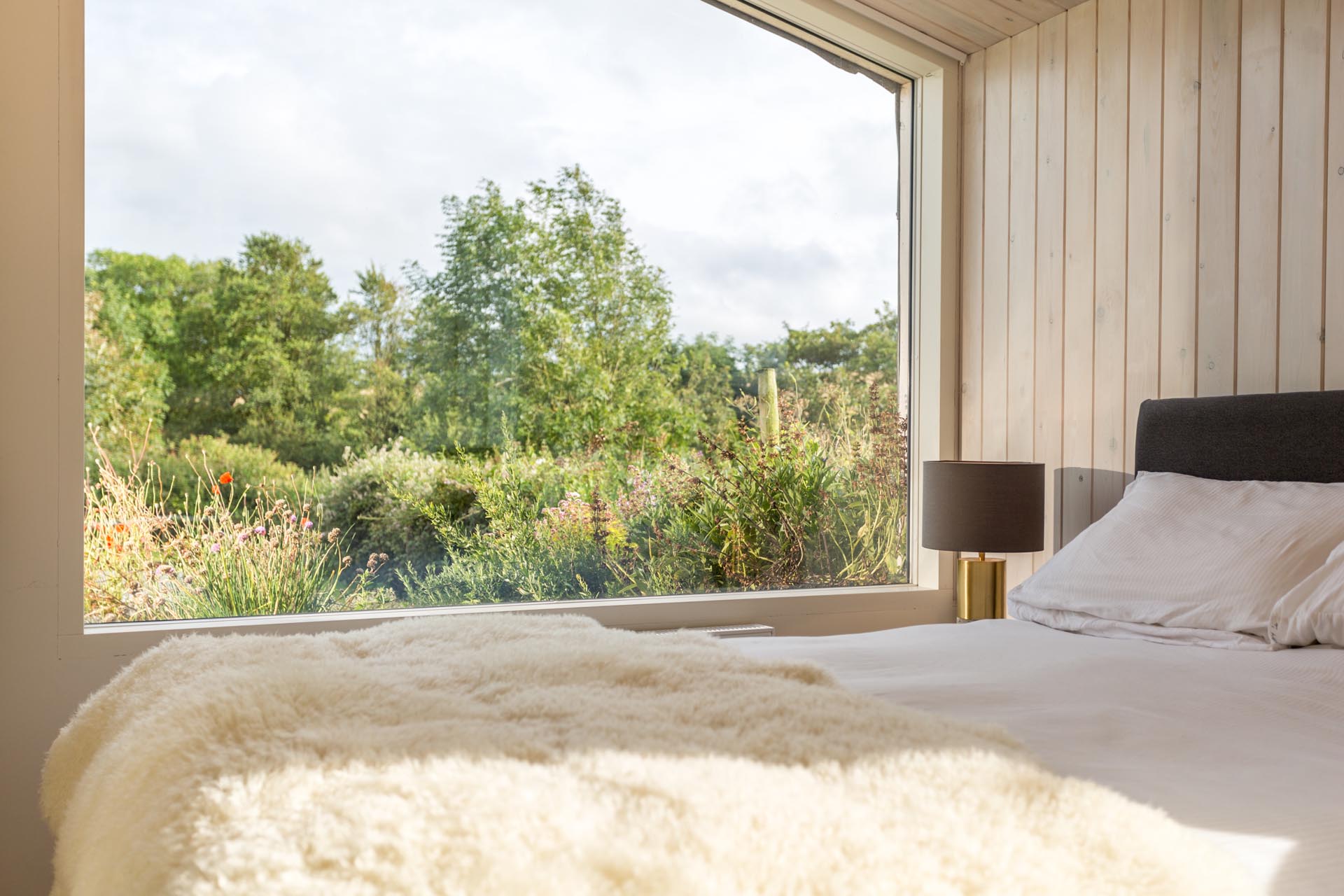 In this modern bedroom, there's wood walls and ceiling, and a large picture window perfectly frames the garden views.