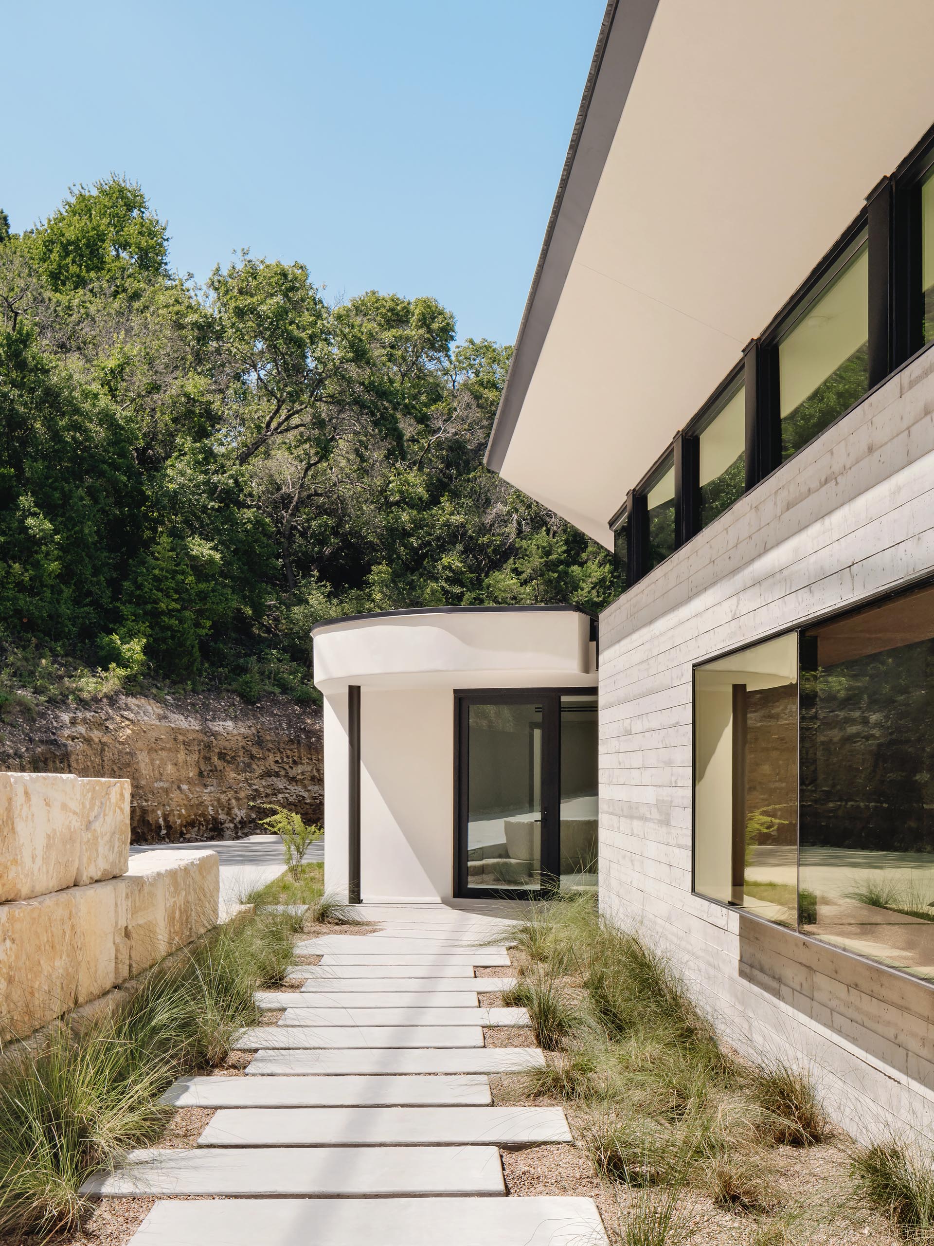 A path lined with grasses runs alongside this modern house and its board formed concrete wall, and guides people to the entryway with a wood accent wall.