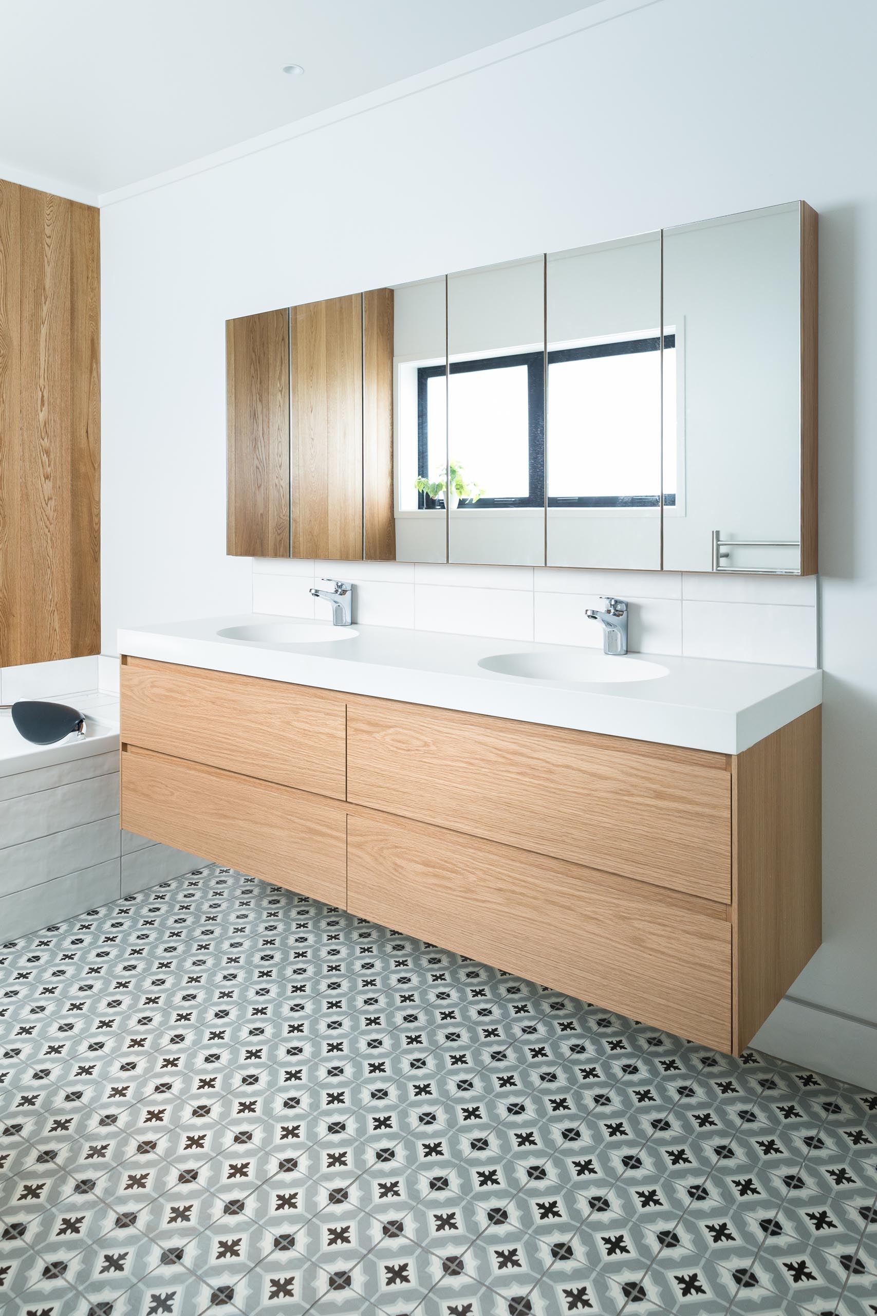 This modern bathroom features a double sink wood vanity, patterned tile flooring, a built-in bathtub, and a wood accent wall.