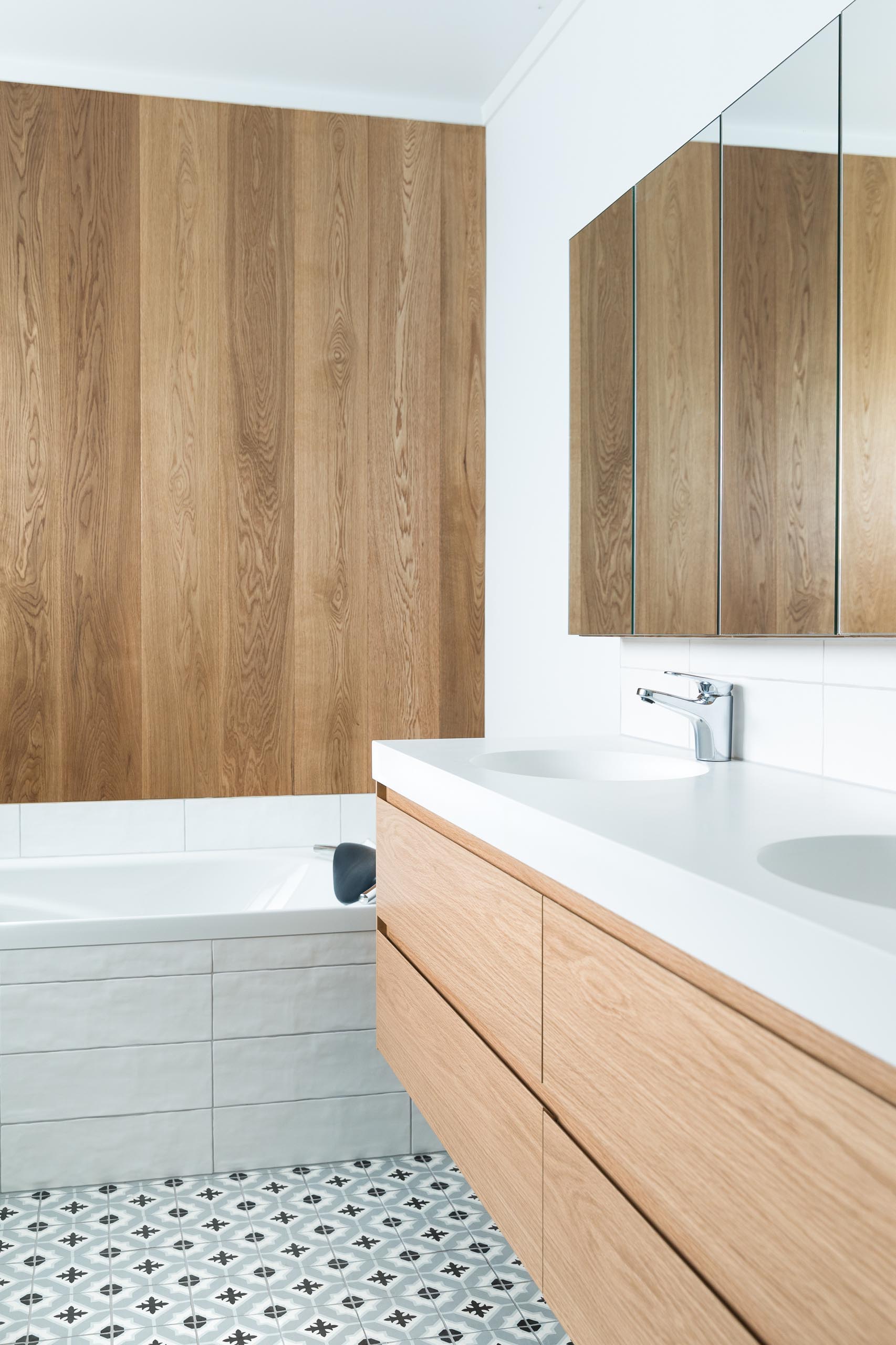 This modern bathroom features a double sink wood vanity, patterned tile flooring, a built-in bathtub, and a wood accent wall.