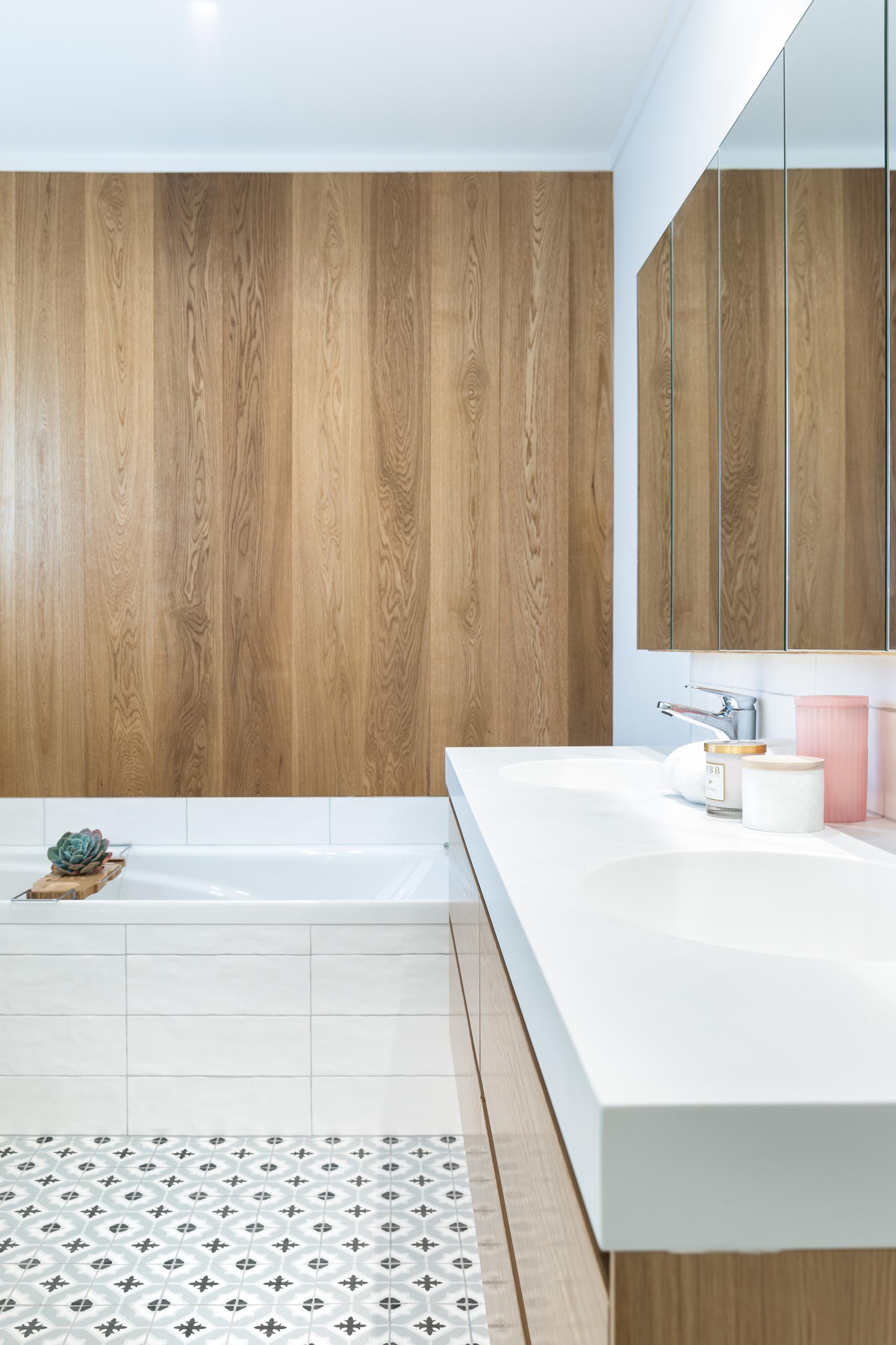 This modern bathroom features a double sink wood vanity, patterned tile flooring, a built-in bathtub, and a wood accent wall.