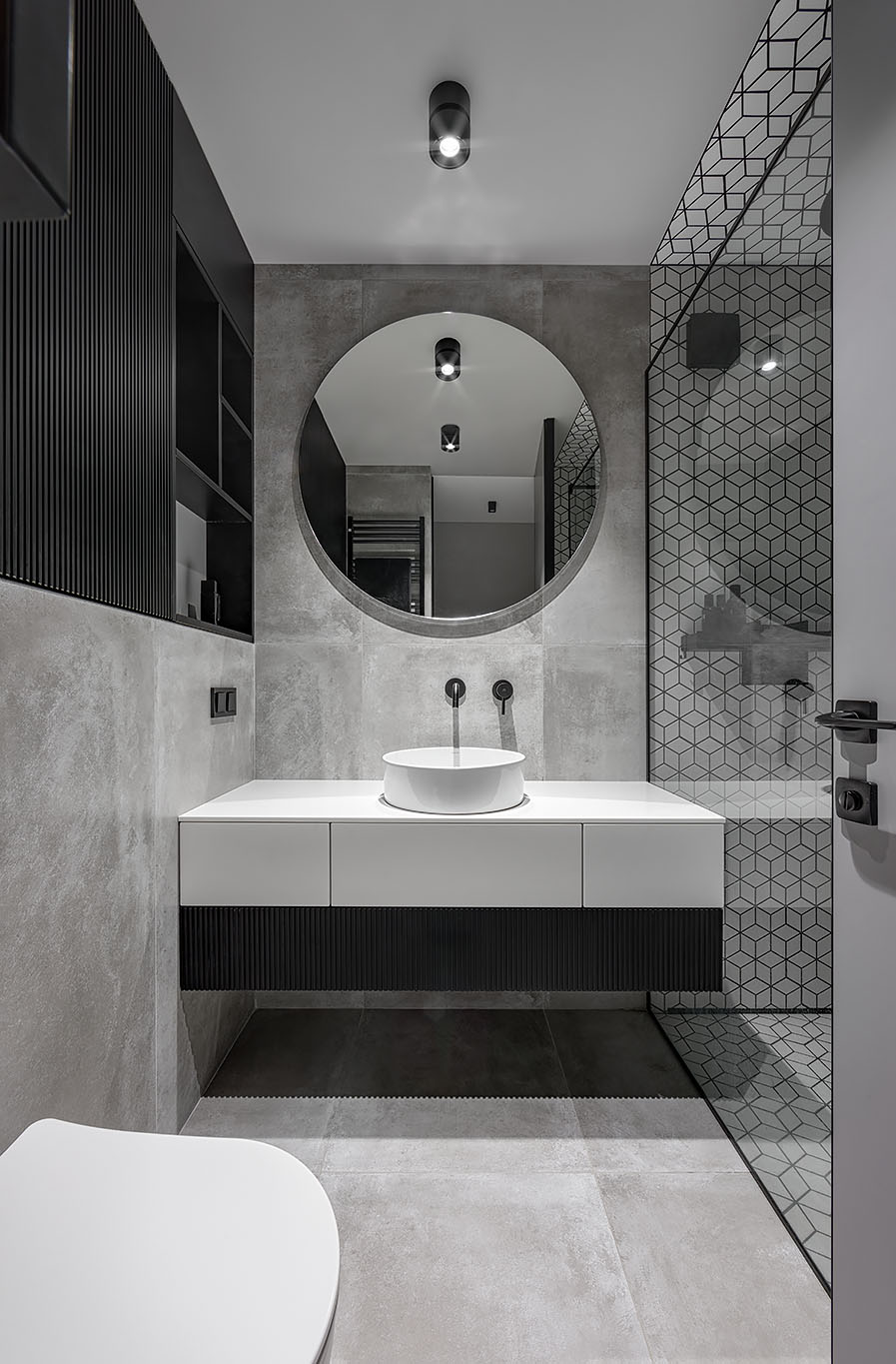 Phòng tắm bao gồm nội thất màu trắng, xám và đen, với bàn trang điểm màu trắng sáng, tường và gạch lát nền bê tông, gạch hình học màu trắng với vữa đen và giá đỡ tùy chỉnh màu đen.