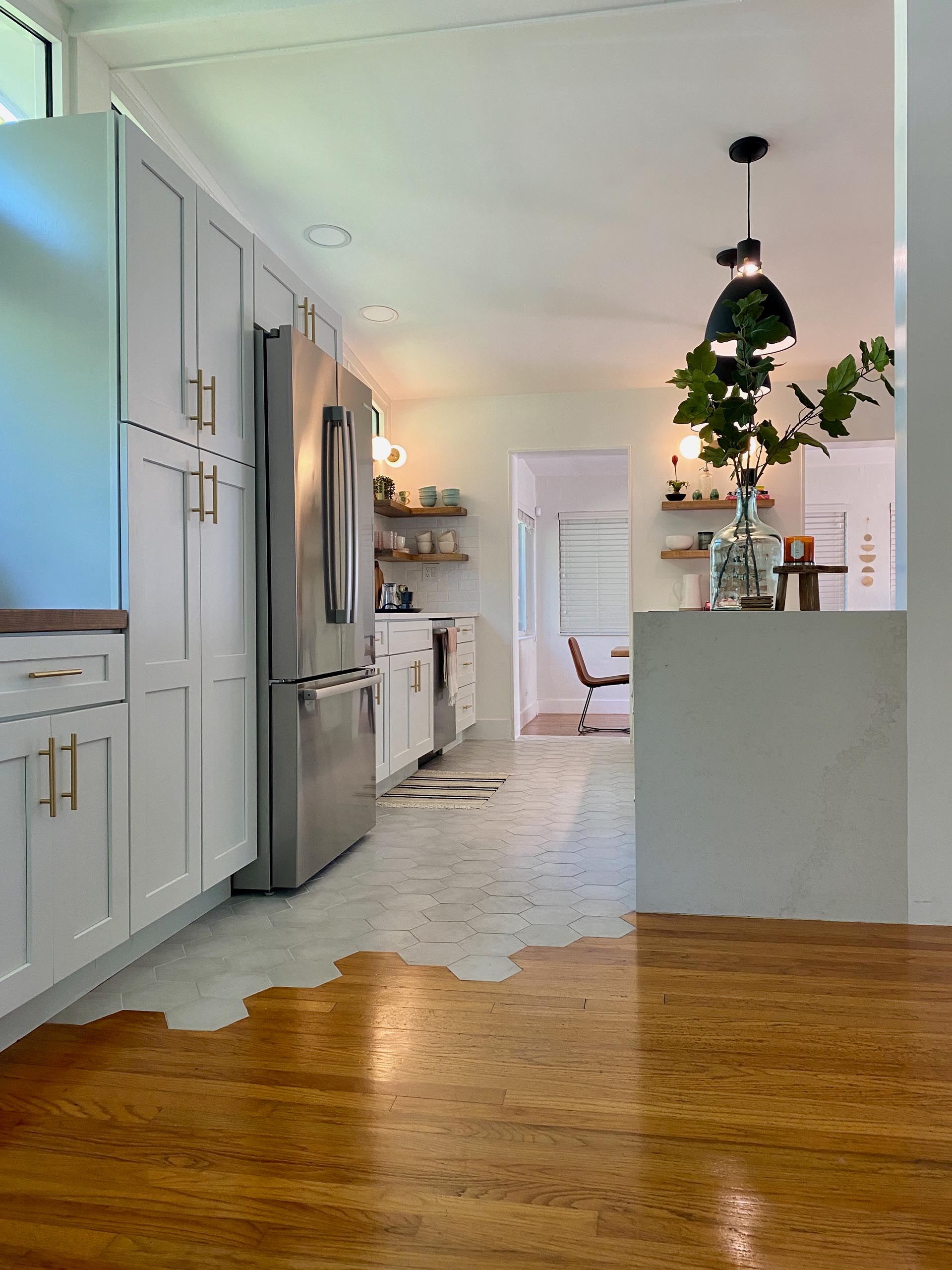 A modern and open kitchen with a peninsula, white cabinets, gold accents, open wood shelving, and hexagonal floor tiles.