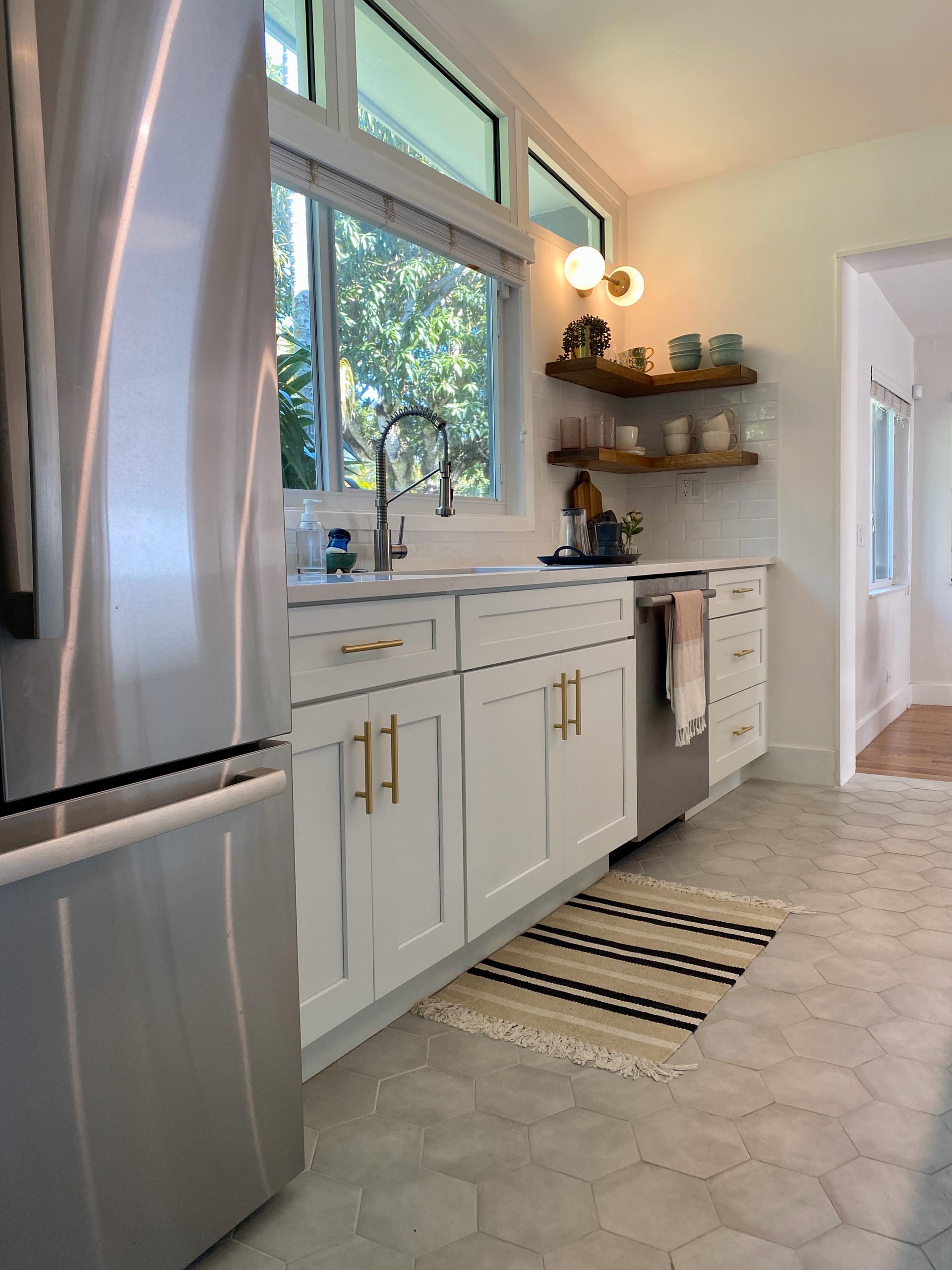 A modern and open kitchen with a peninsula, white cabinets, gold accents, floating wood shelving, and hexagonal floor tiles.