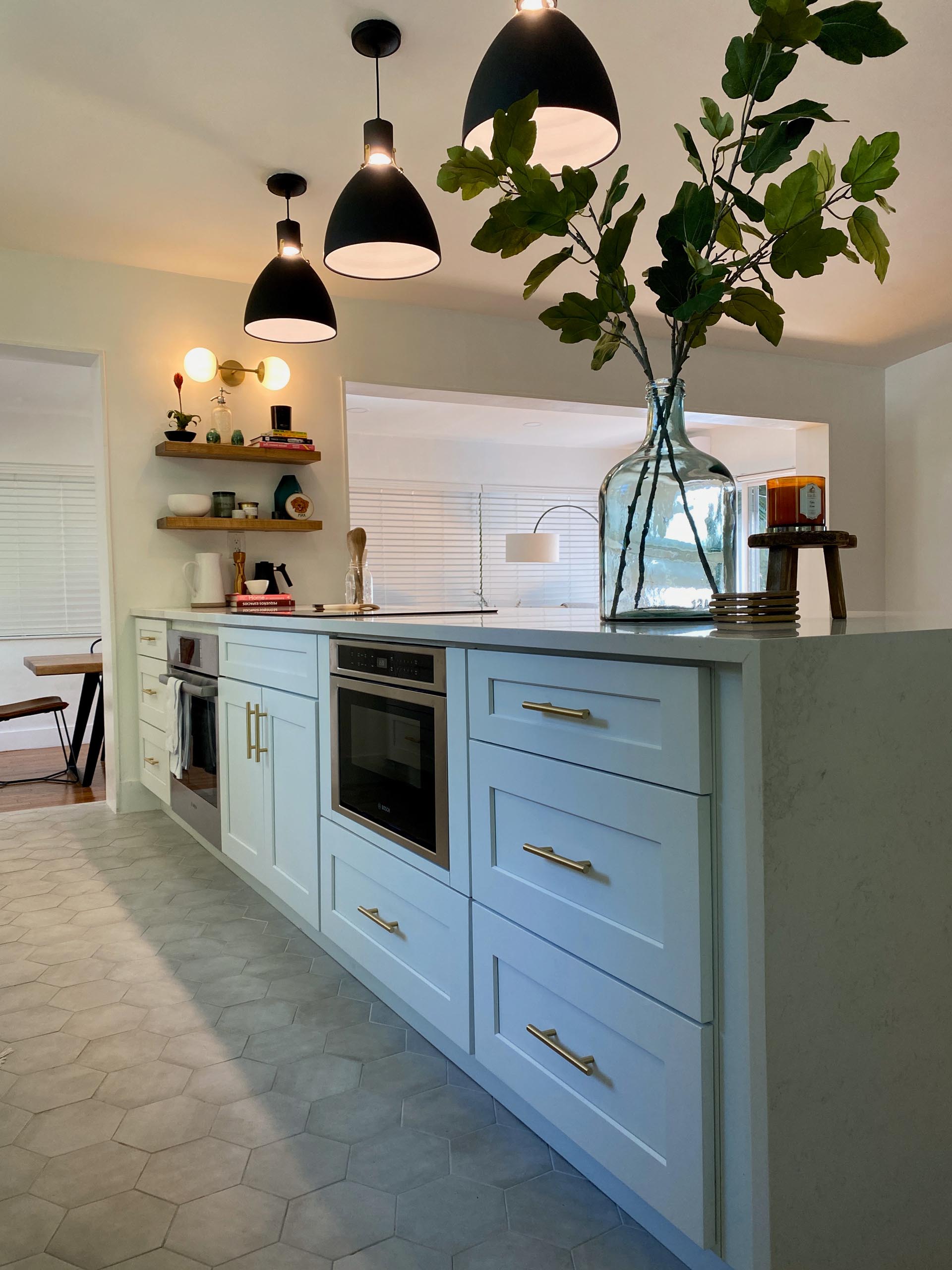 A modern and open kitchen with a peninsula, white cabinets, gold accents, floating wood shelving, and hexagonal floor tiles.