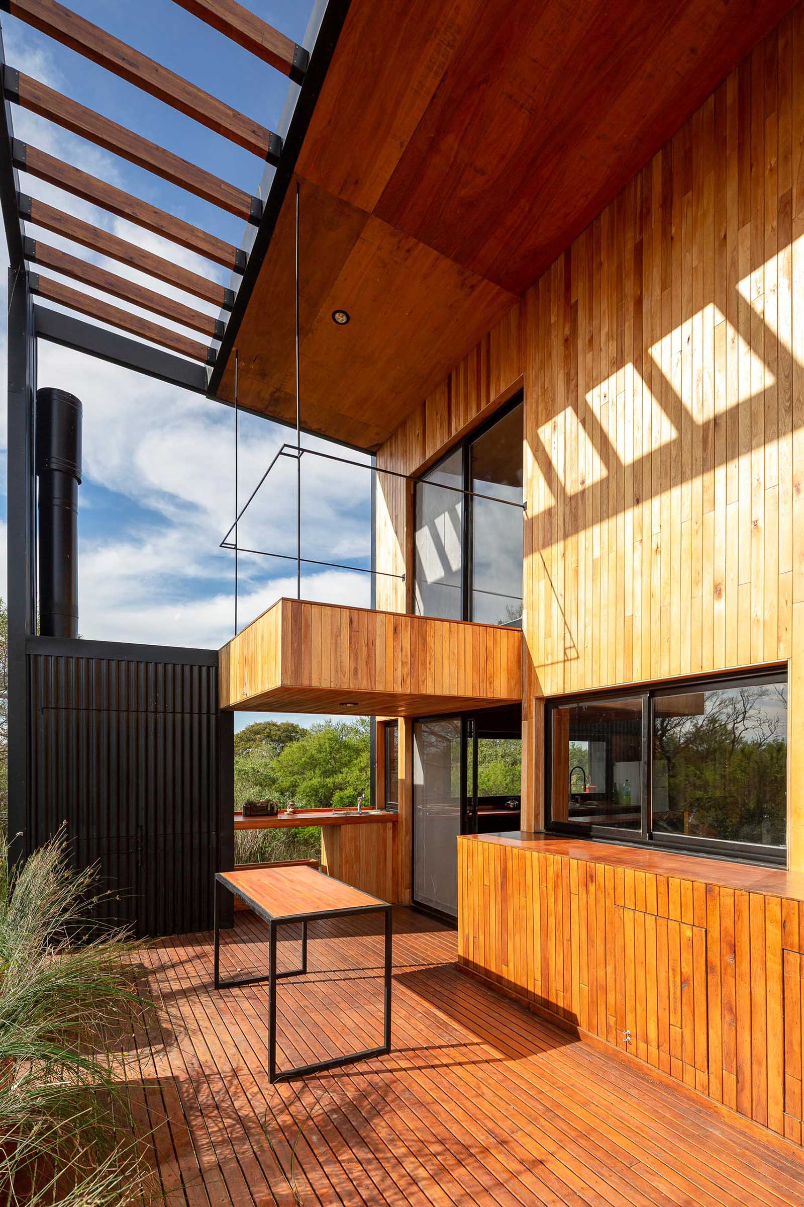 Stairs connect the ground level to the deck of this small house, includes matching wood planters, outdoor dining area, and a small kitchen space with a sink.