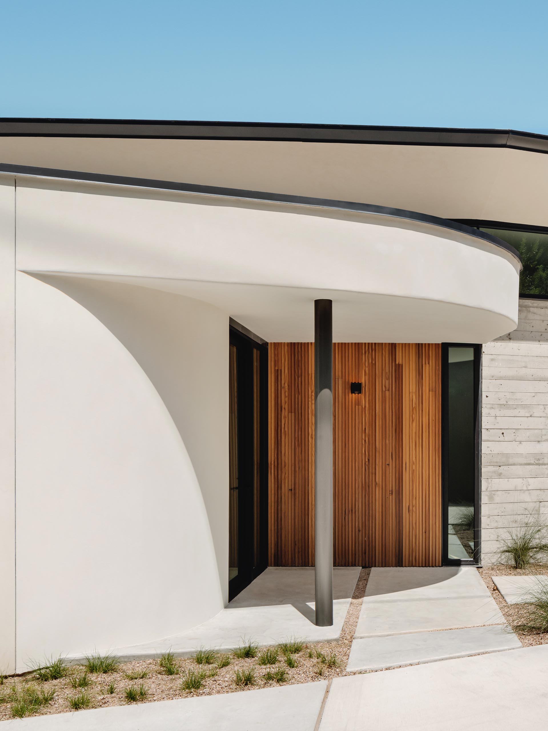 A path lined with grasses runs alongside this modern house and its board formed concrete wall, and guides people to the entryway with a wood accent wall.