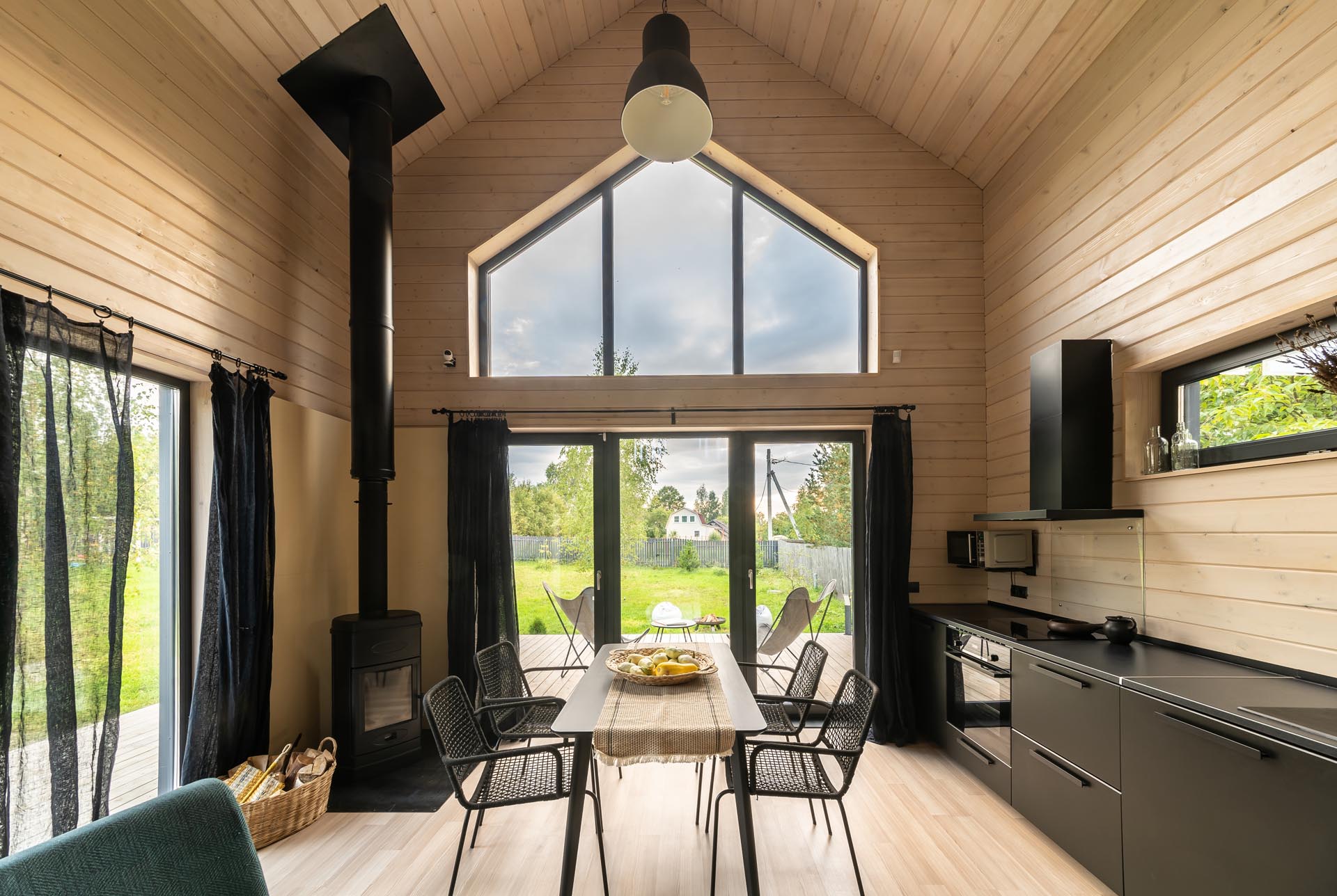 A modern barn-inspired open plan dining area and kitchen with tongue and groove wood siding and black accents, like the kitchen cabinets, the fireplace, the curtains, and furniture.