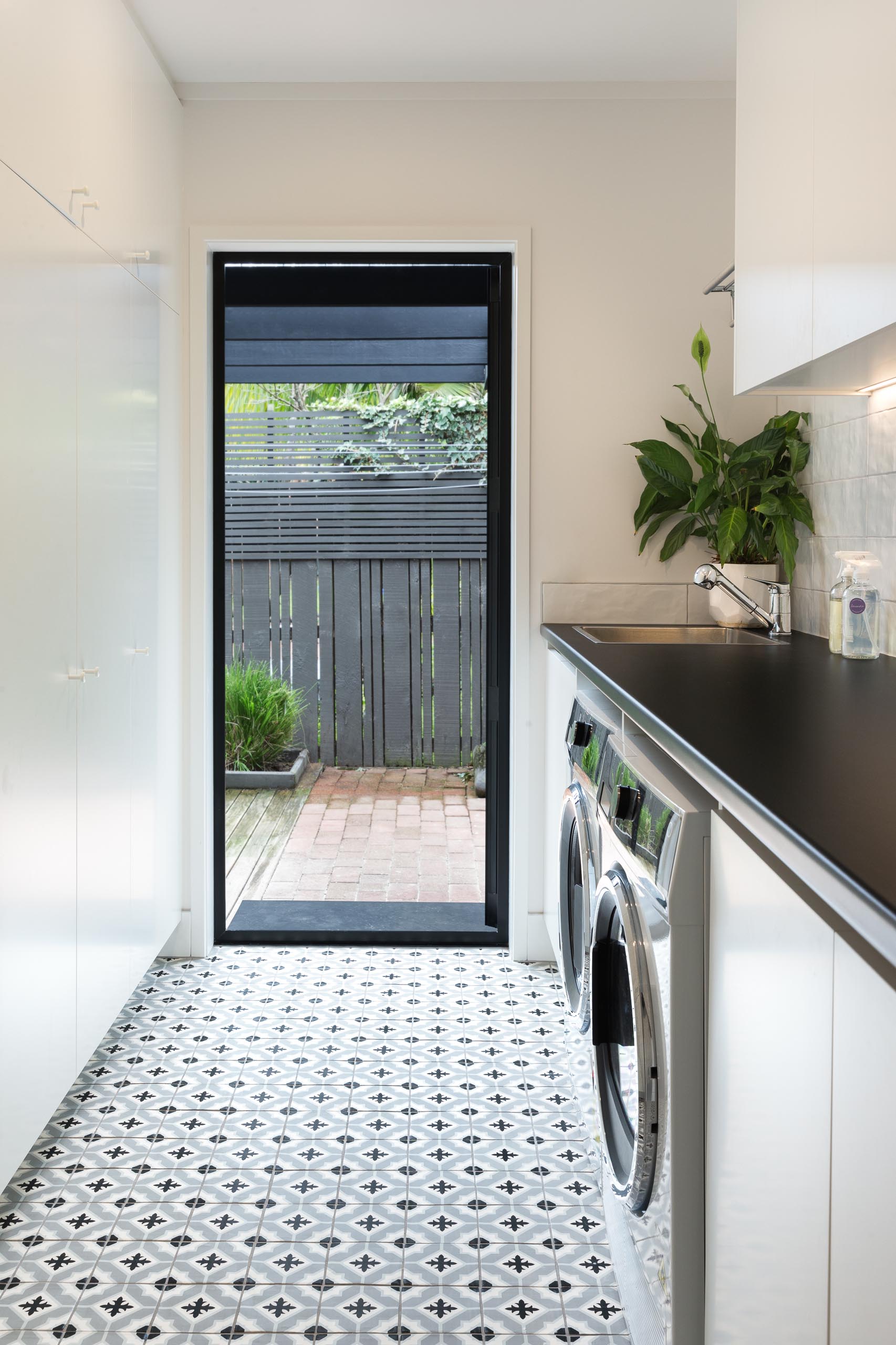 This modern laundry room has been designed with patterned floor tiles, floor-to-ceiling cabinets that add plenty of storage, and a black countertop travels across both the washing machine and dryer, and complements the black door frame.