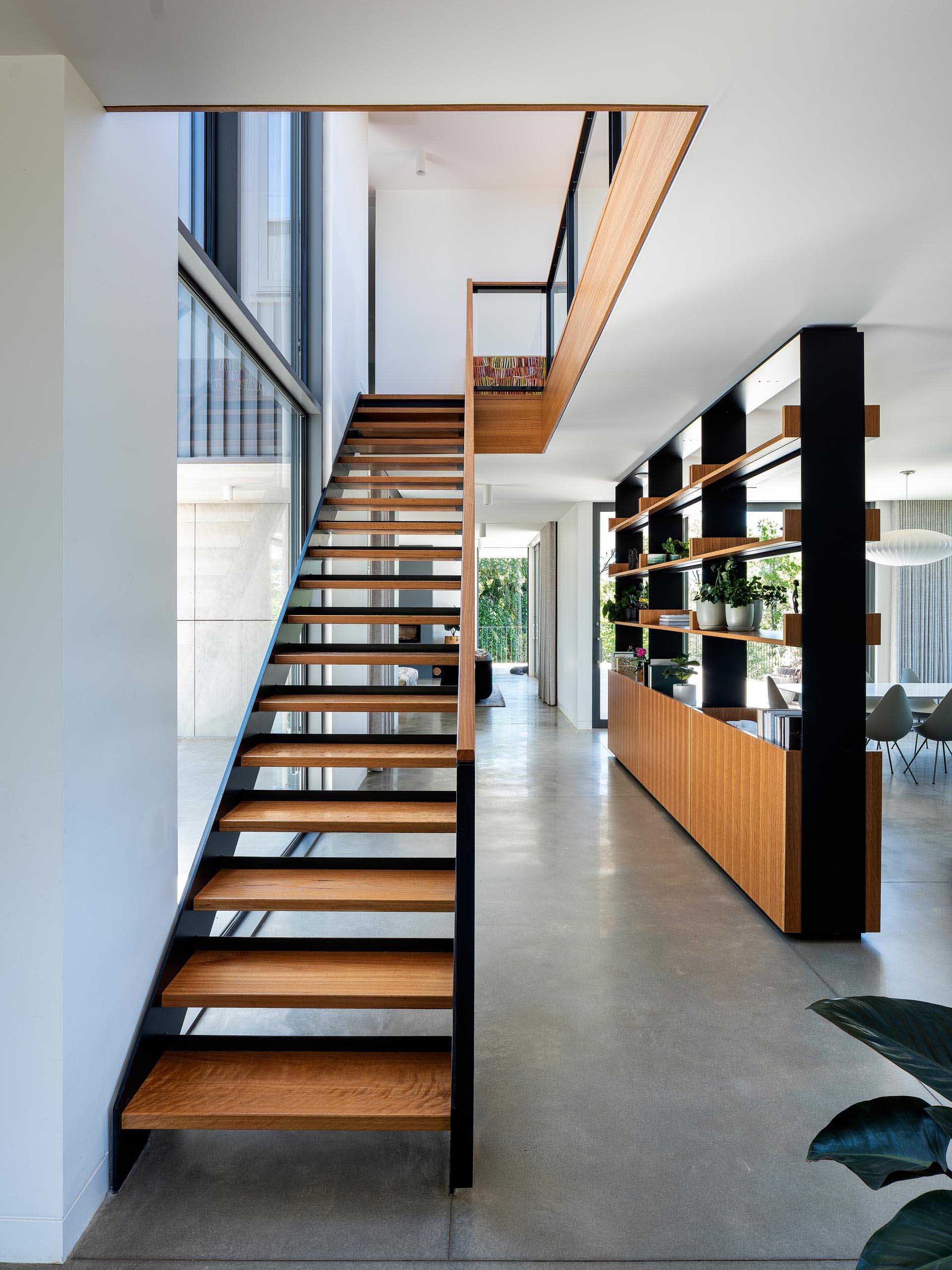 The shelving unit, with its black metal frame and wood accents, matches the design of the adjacent stairs, and contrasts other materials found throughout the home, like the heated concrete floors.