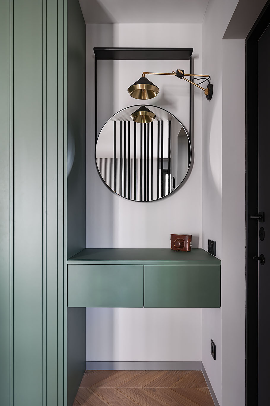 A small nook has been filled with a custom-built floating storage cabinet that also acts as an entryway table. Above, there's a round mirror, while a wall-mounted lamp adds light.