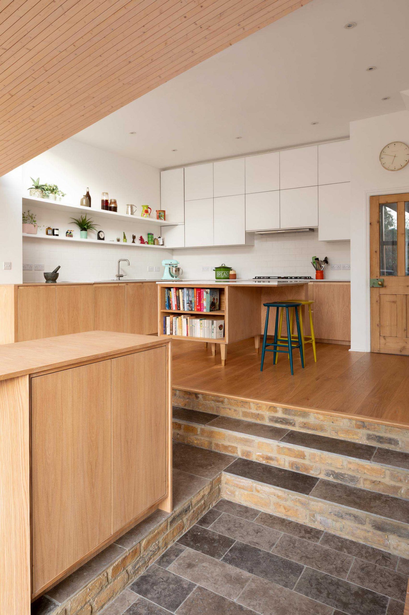 A contemporary kitchen with minimalist white cabinets that fill the wall, open white shelving, a small island with recipe book storage, and lower wood cabinets.