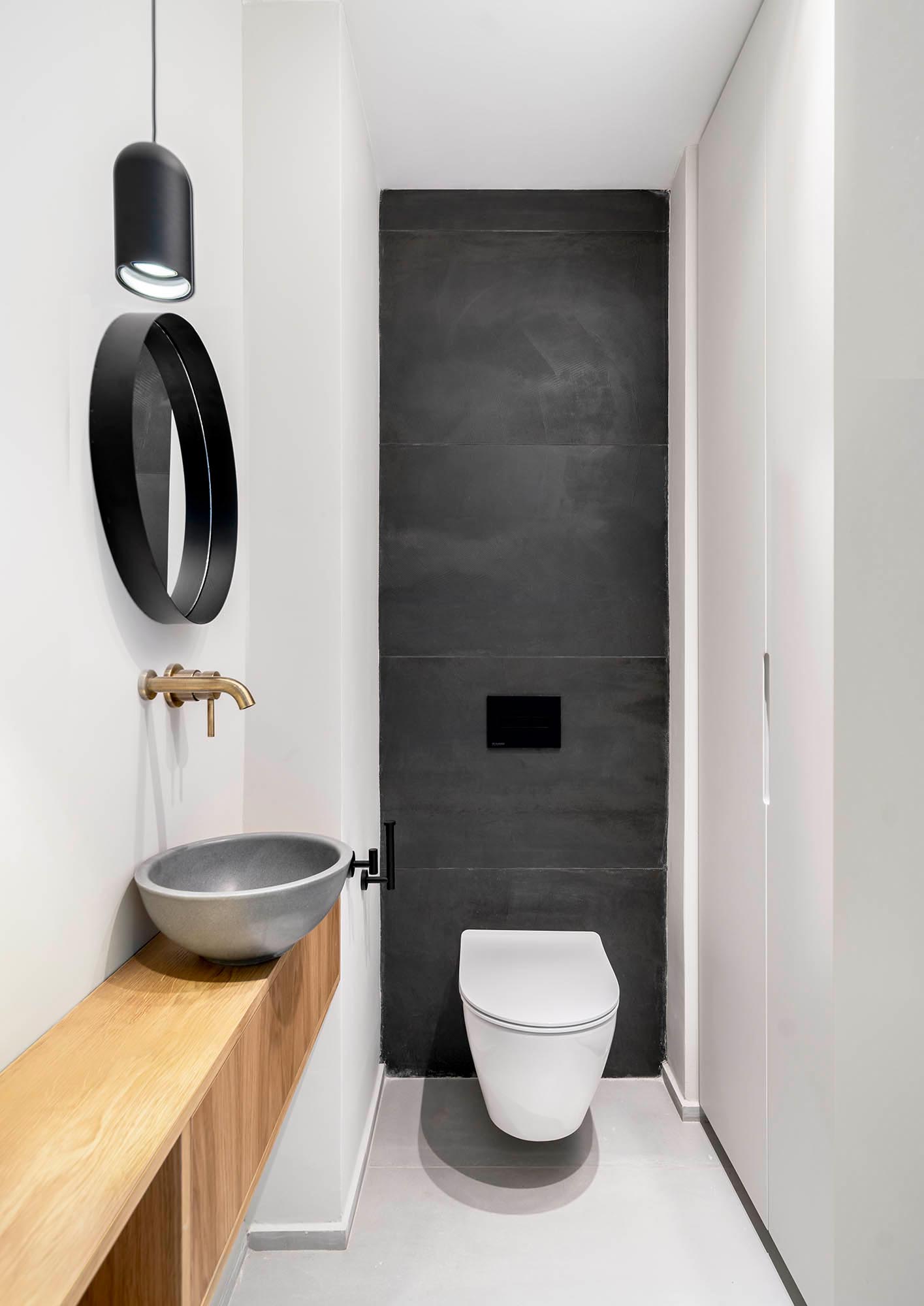 A powder room with large format matte black tiles line the wall behind the toilet, a wood vanity topped with a gray vessel sink, and bronze hardware.