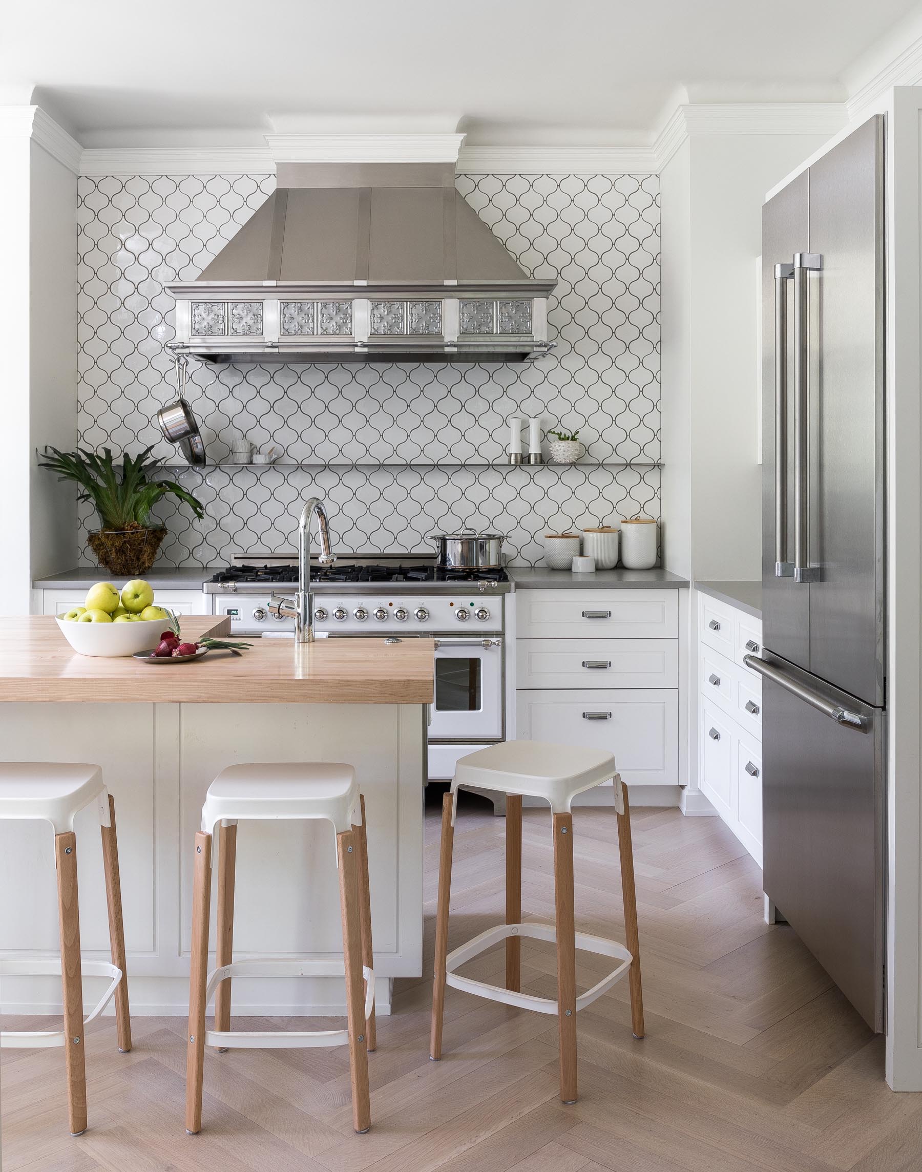 In this remodeled kitchen, there's wood flooring in a herringbone pattern, white cabinets, a patterned white tile with dark grout, and stainless steel appliances. An island with a wood countertop complements the flooring.