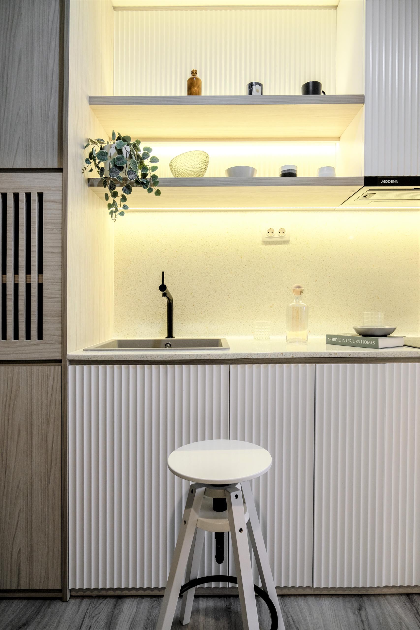 The kitchen of a small apartment includes textured cabinet fronts that help to define the kitchen area, while added lighting underneath the shelves helps brighten the countertops.
