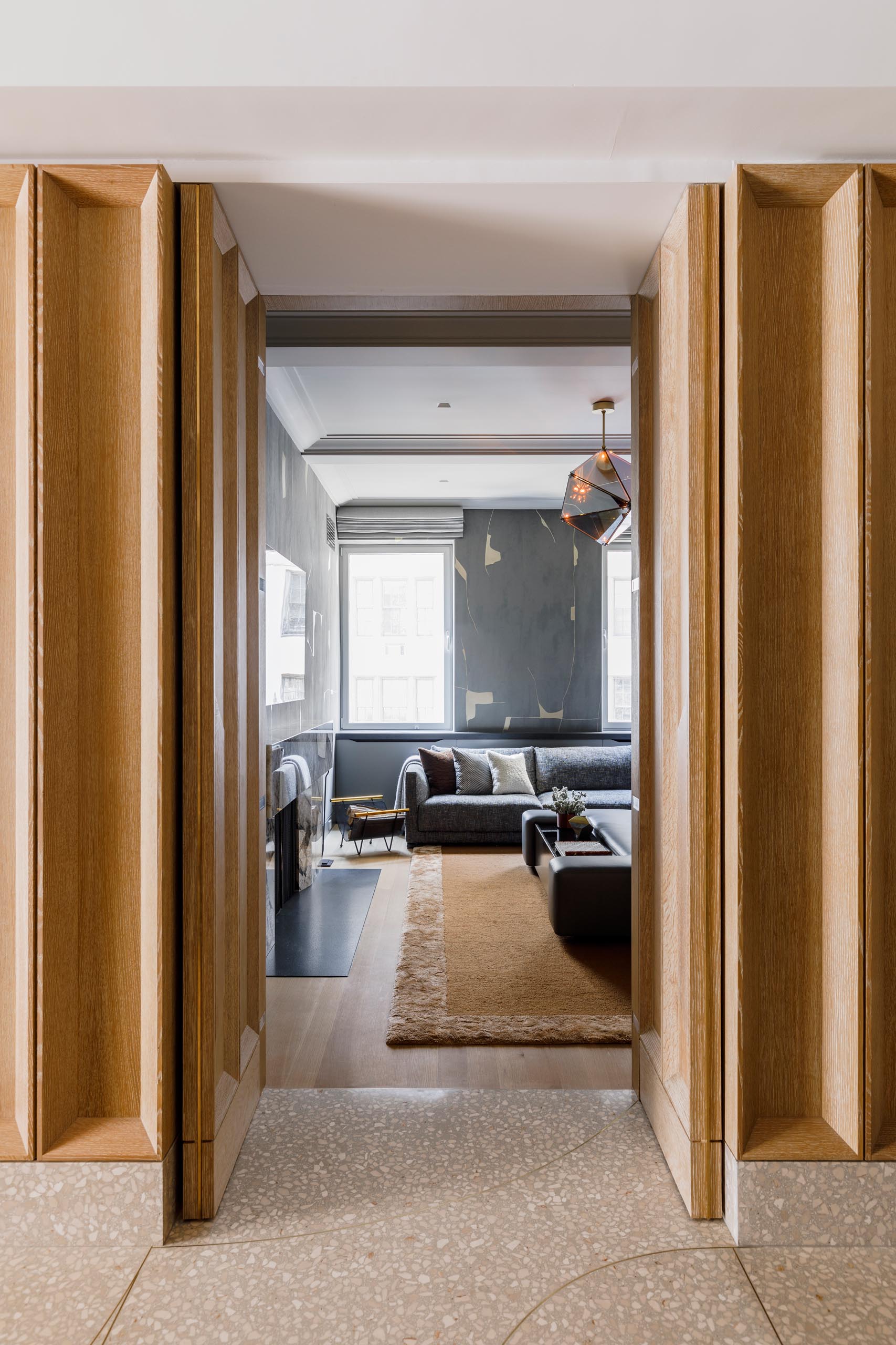 In this modern media room, there's hand painted “Kintsugi” wallcovering by Porter Teleo, a Patagonia marble fireplace, a custom leather, walnut and glass ottoman by MKCA and fabricated by Manzanares Furniture, a sofa by Poliform, and an overhead “Maxihedron” pendant by Bec Brittain for Roll and Hill.