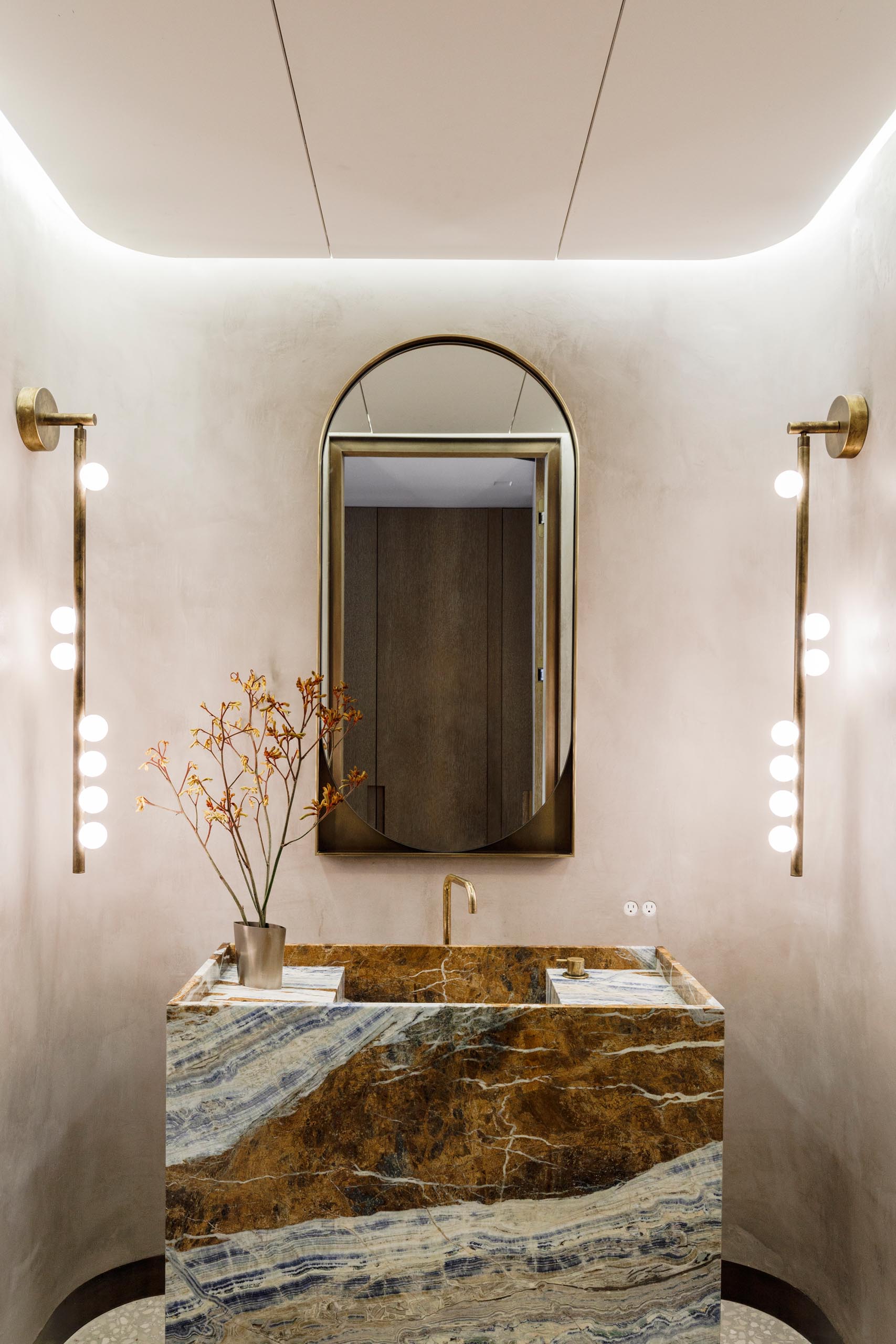 This powder room includes a stone vanity in figural Cassiopeia marble, curved venetian plaster walls with LED cove lighting, sconces by Lindsey Adelman, a custom bronze mirror by Kin and Company, and a faucet by Vola.