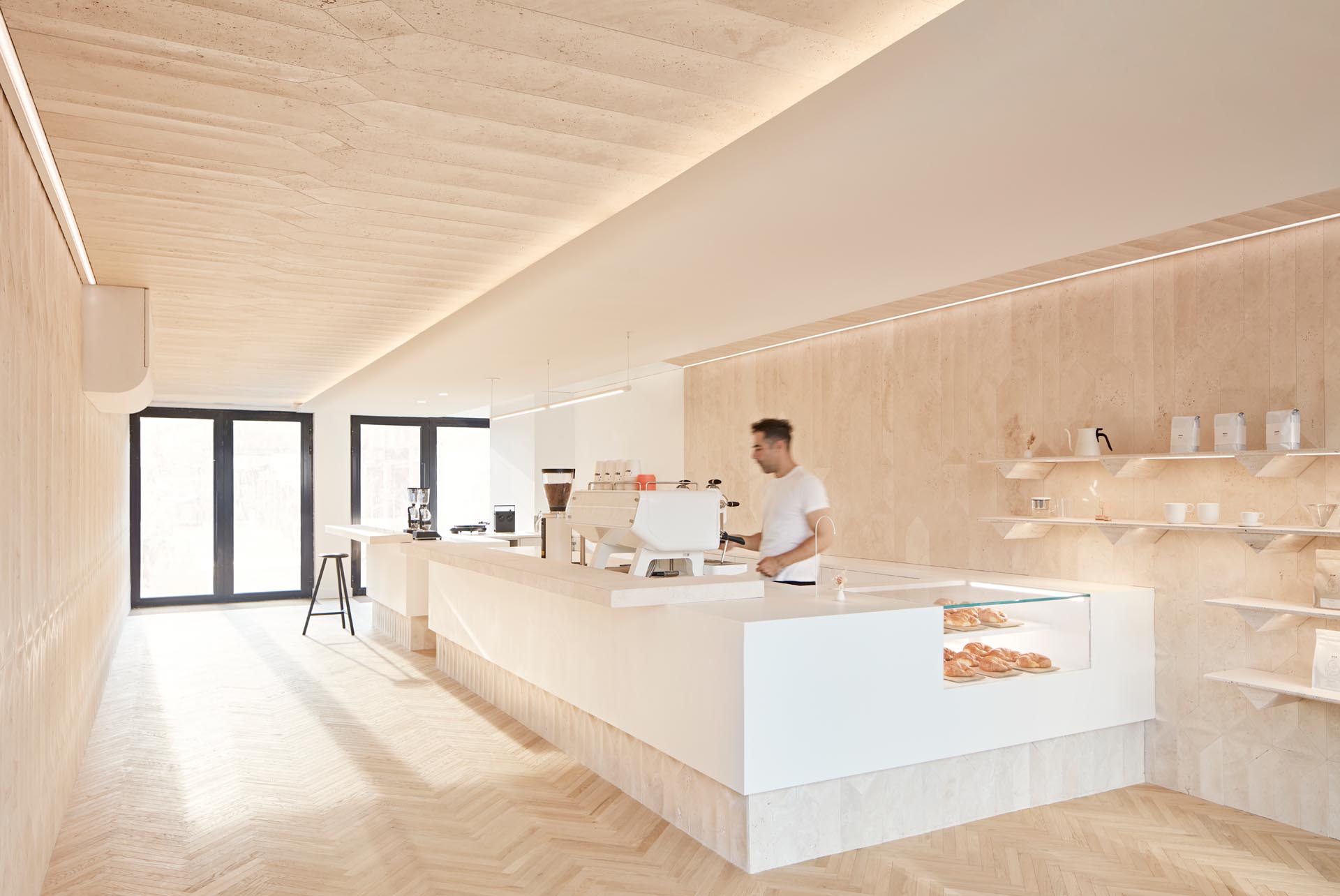 A modern coffee shop inside a shipping container has a large bar, herringbone wood floors, and travertine walls.