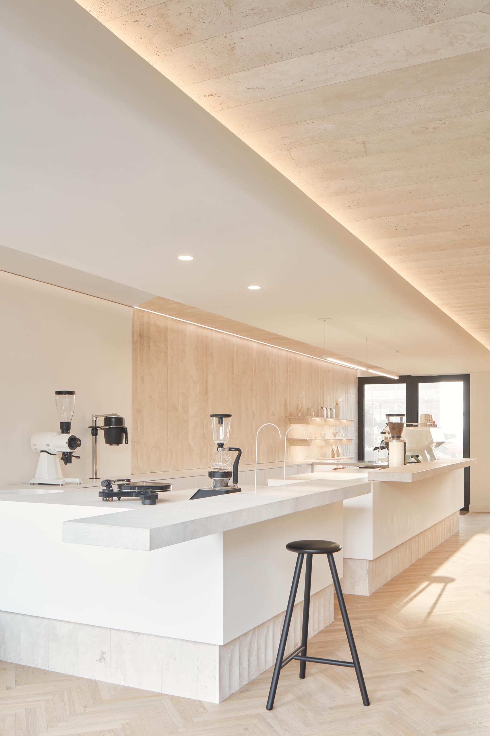 A modern coffee shop inside a shipping container has a large bar, herringbone wood floors, and travertine walls.