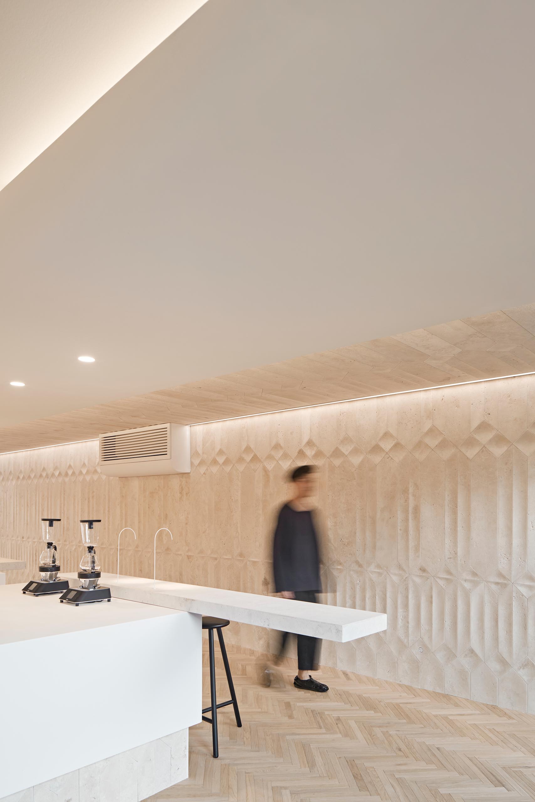 A modern coffee shop inside a shipping container has a large bar, herringbone wood floors, and travertine walls.