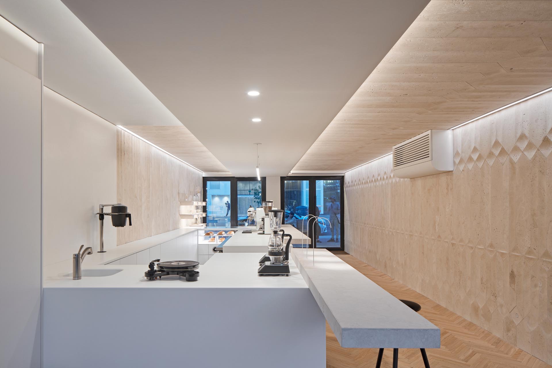 A modern coffee shop inside a shipping container has a large bar, herringbone wood floors, and travertine walls.