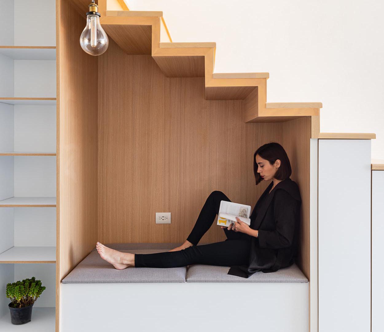 An under-stair wood lined nook has bench seating with an upholstered cushion, as well as an outlet and a nearby pendant light. To the left of the nook is an open shelving unit, while to the right, are small cabinets.