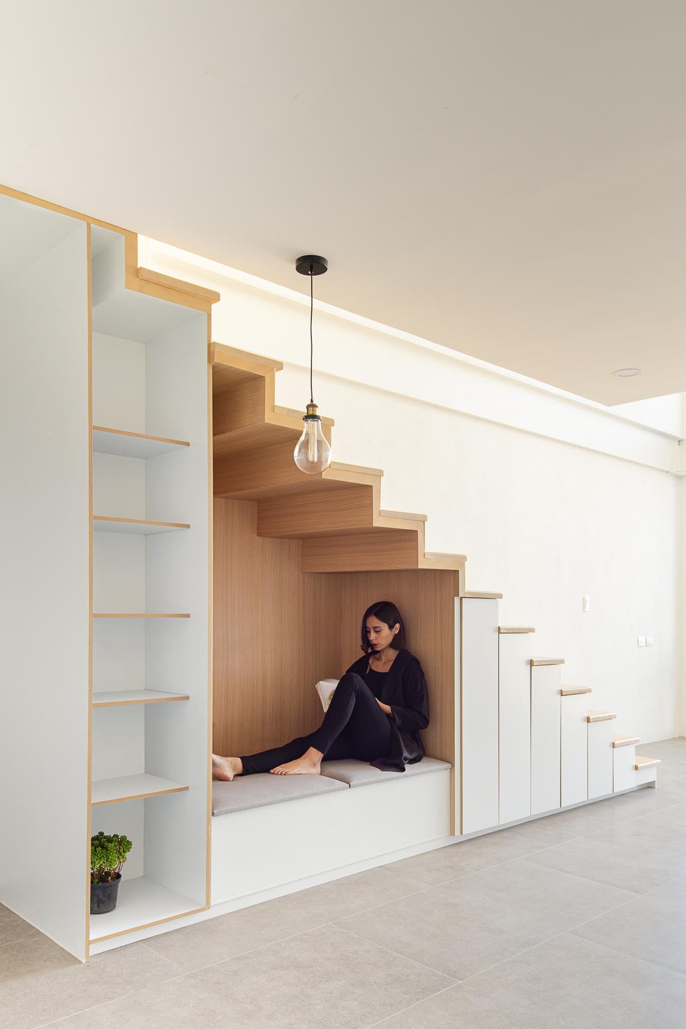 An under-stair wood lined nook has bench seating with an upholstered cushion, as well as an outlet and a nearby pendant light. To the left of the nook is an open shelving unit, while to the right, are small cabinets.