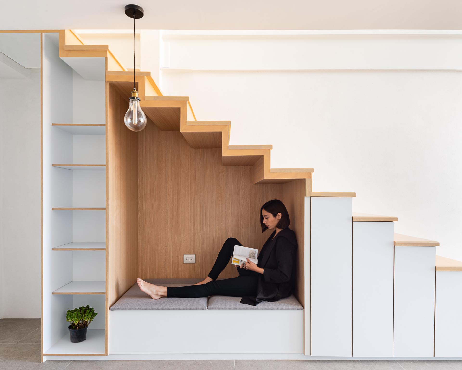 An under-stair wood lined nook has bench seating with an upholstered cushion, as well as an outlet and a nearby pendant light. To the left of the nook is an open shelving unit, while to the right, are small cabinets.