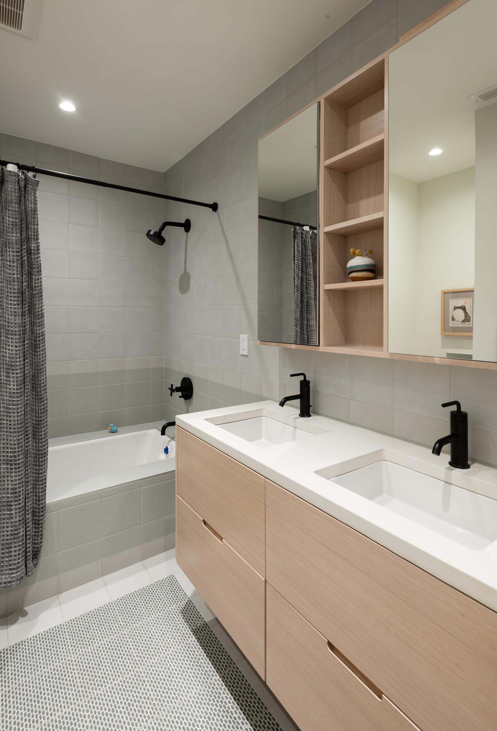 A modern bathroom with a light wood vanity, white countertop, and a shower/bath lined with square gray tiles.