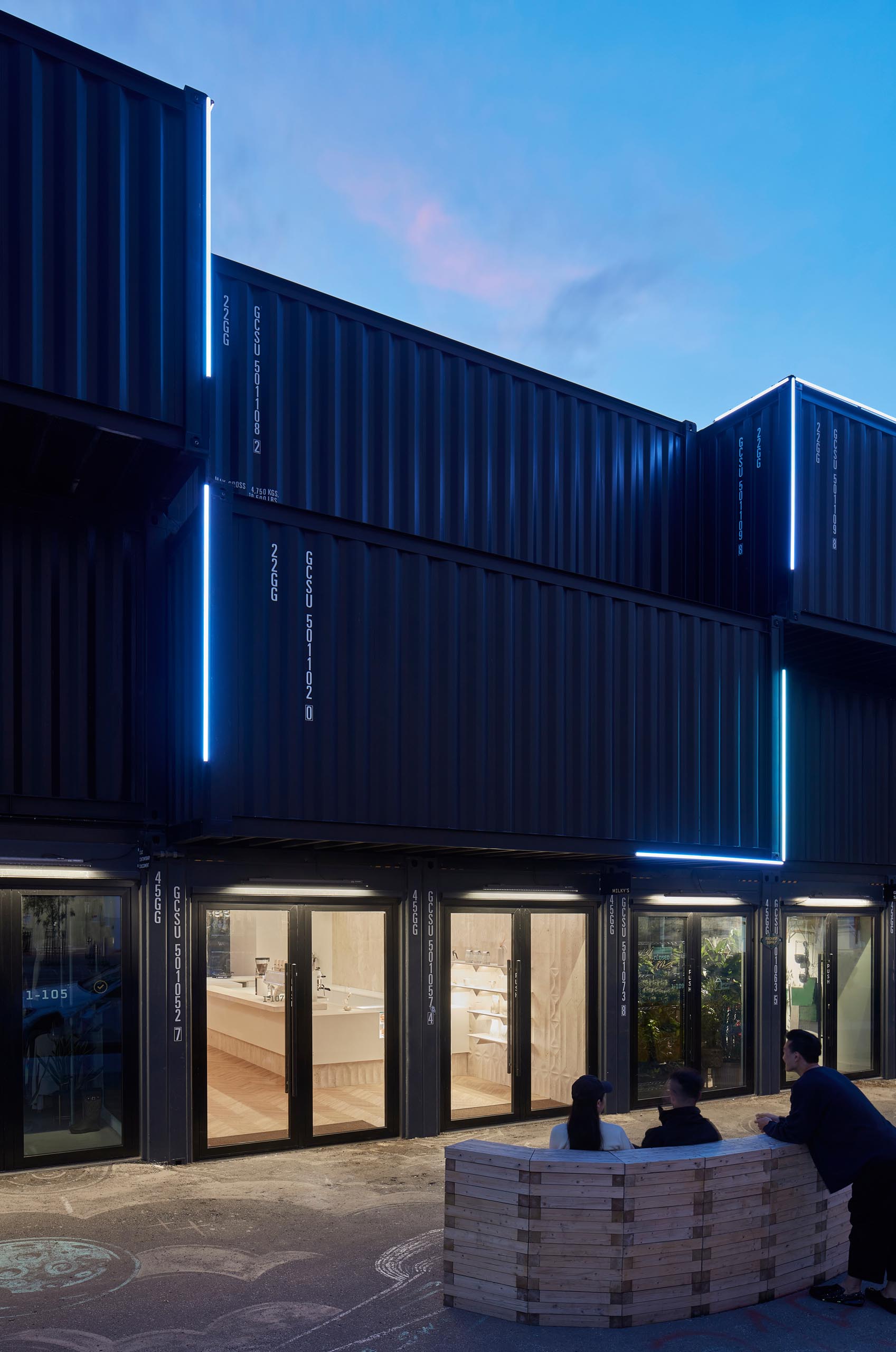 A modern coffee shop inside a shipping container has a large bar, herringbone wood floors, and travertine walls.