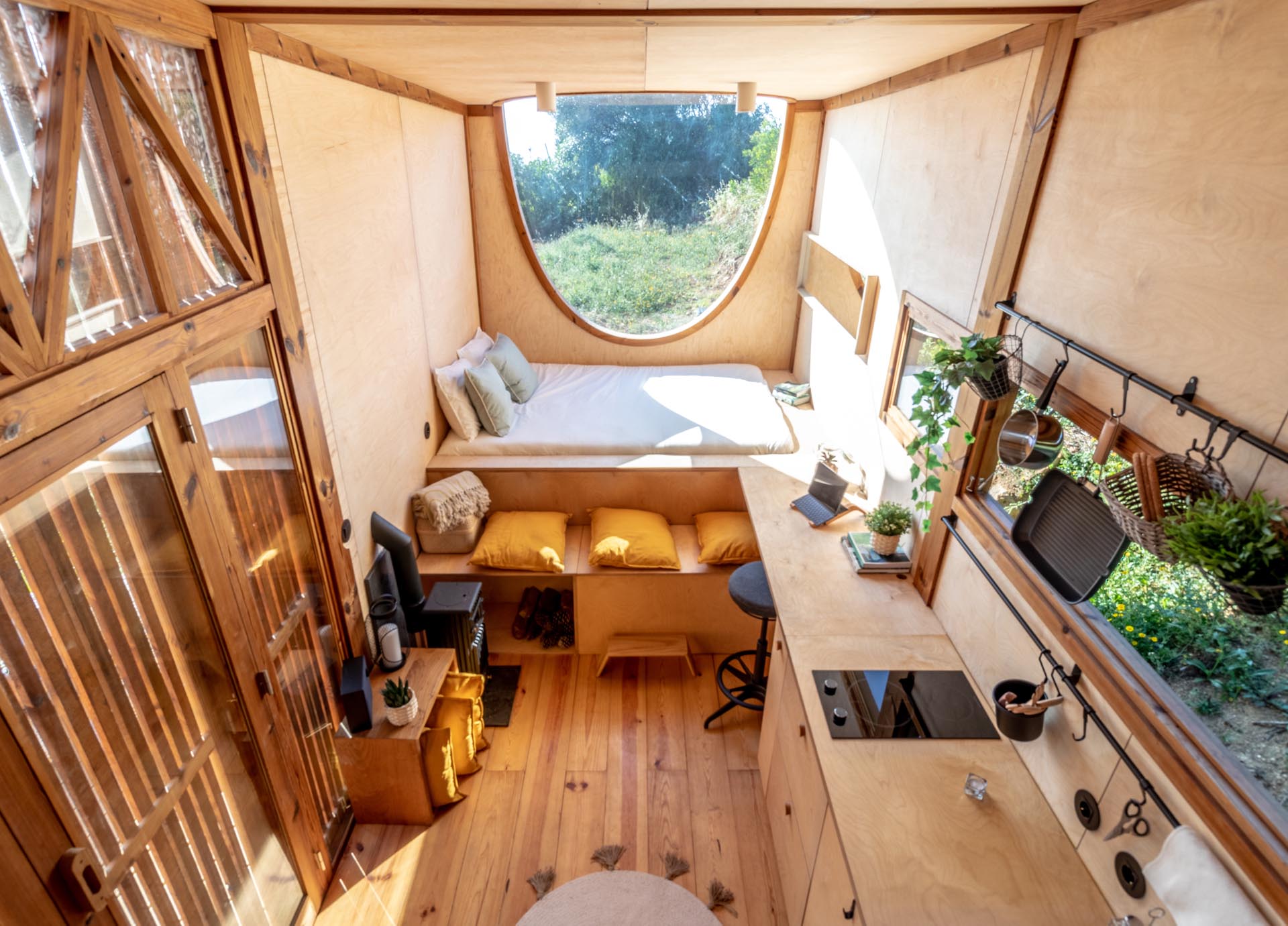 This modern tiny house includes a kitchen, an oval window, a small desk / dining area, a bench, and a bed.