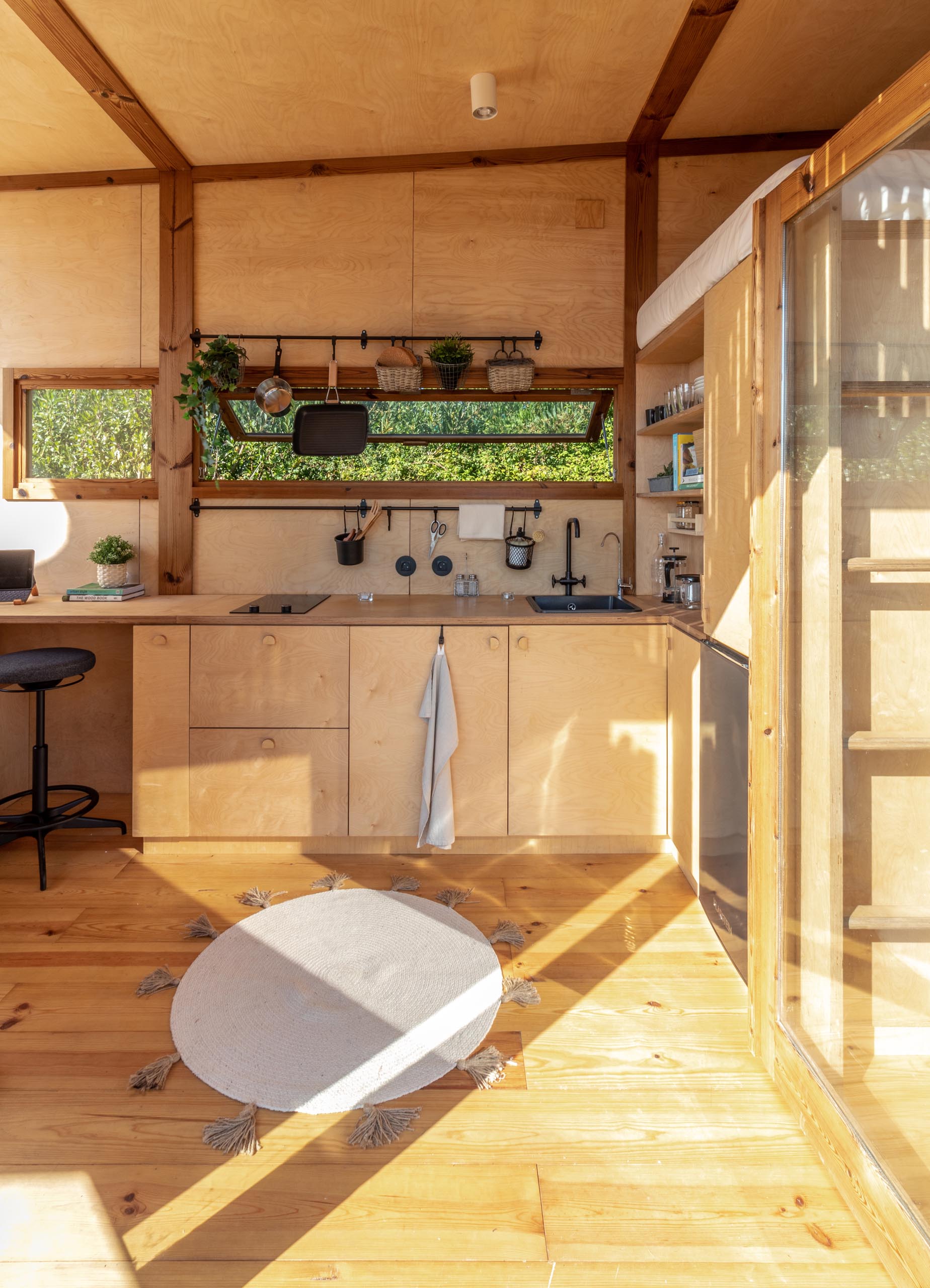Stepping inside this modern tiny house, and the interior walls and ceiling are covered with birch plywood panels. A small kitchen with a window has a fridge, cooktop, and sink.