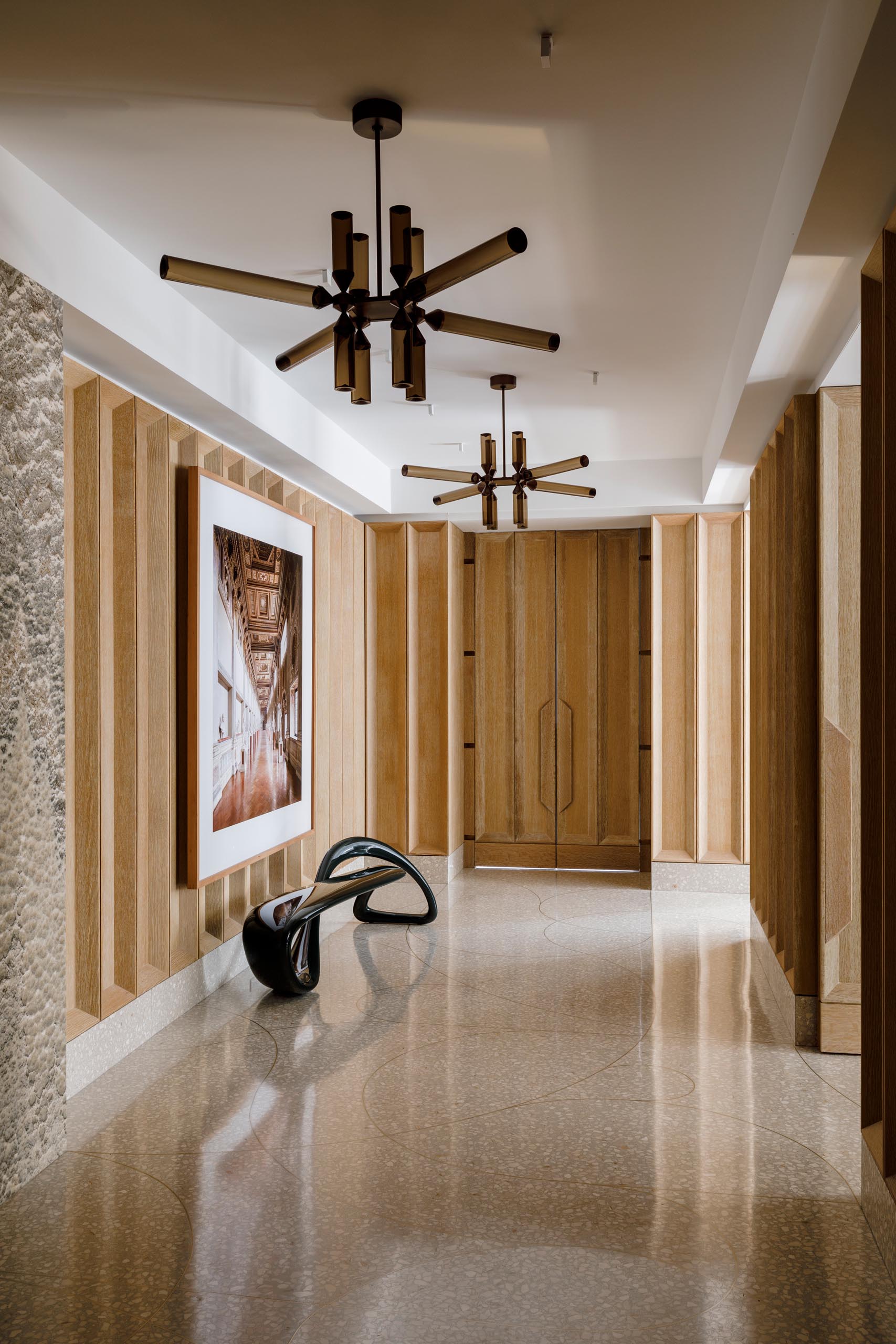 This entryway and gallery includes deep coffered paneling in cerused white oak, custom terrazzo floor with hand bent brass inlay, two “Castle” pendants by Jason Miller for Roll and Hill, photo artwork by Candida Hofer, and a fiberglass bench by Brodie Neill.