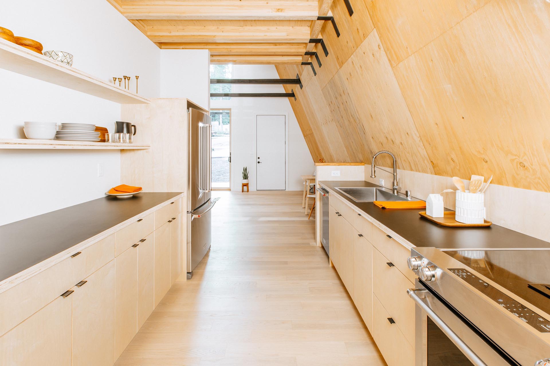 This minimal kitchen in an A-frame cabin includes includes light birch cabinets, minimalist shelving, and dark countertops.