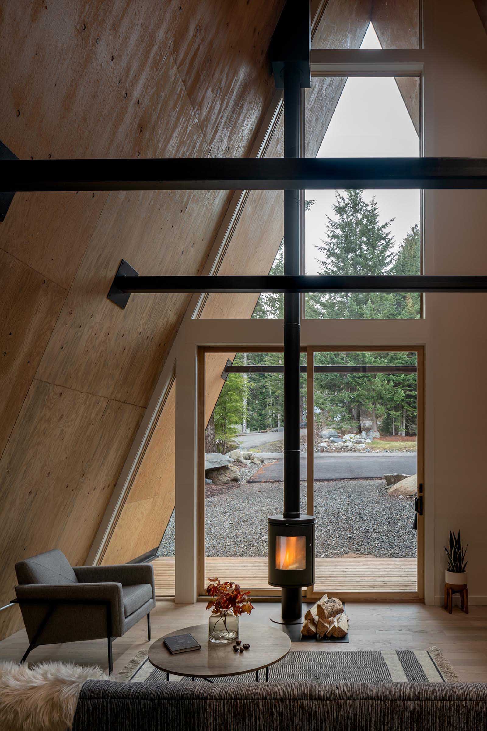 The cozy wood-lined interior of this A-frame cabin includes a fireplace with a black finish that complements the other metal elements. Sliding glass doors open to covered verandahs.