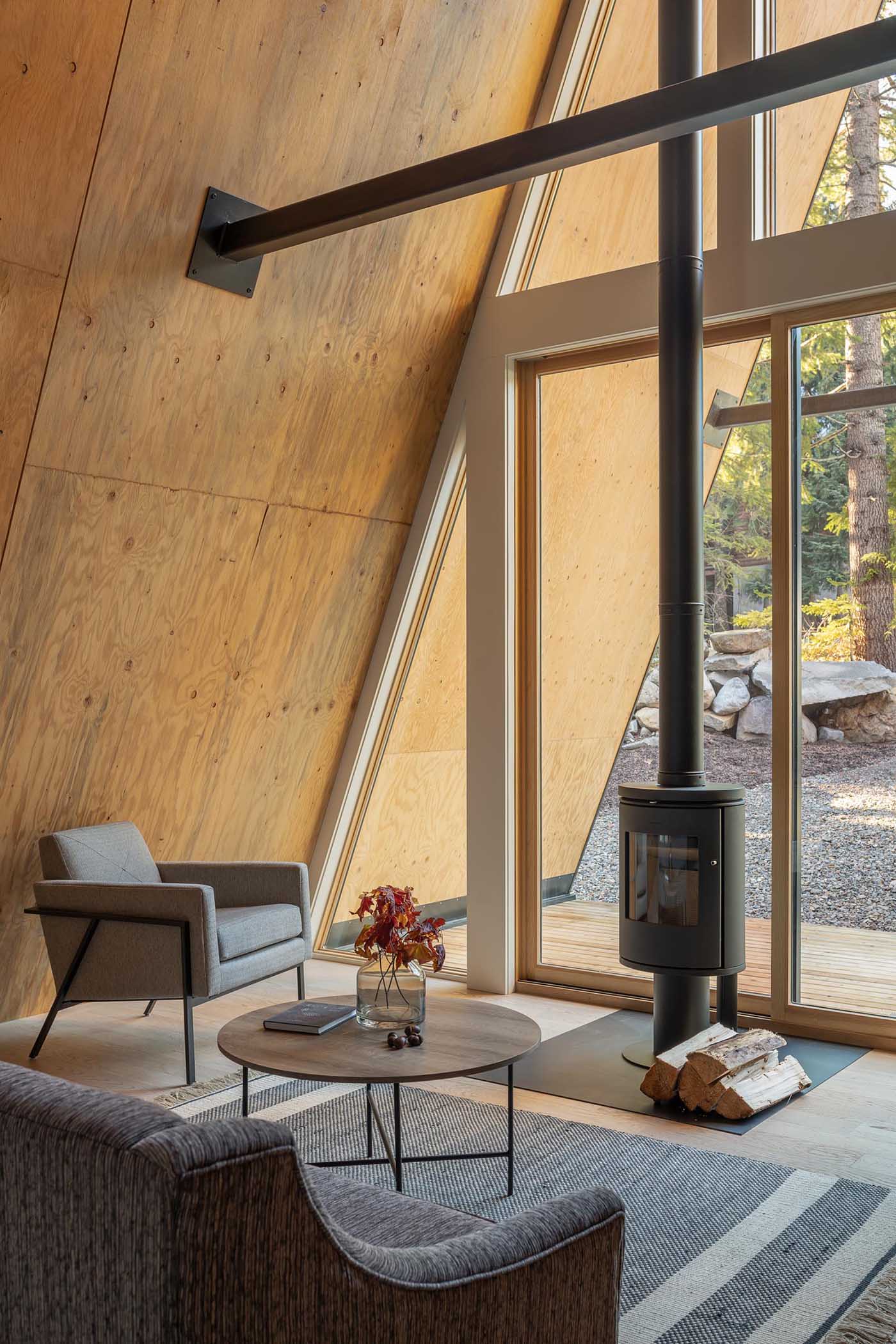 The cozy wood-lined interior of this A-frame cabin includes a fireplace with a black finish that complements the other metal elements. Sliding glass doors open to covered verandahs.