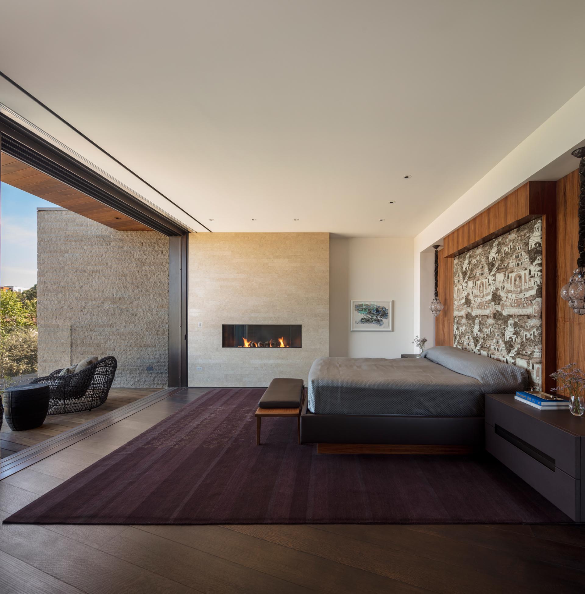 A modern bedroom includes a wood accent wall with an upholstered headboard, a linear fireplace, dark wood floors, and a wall that opens to a deck for views of the valley.
